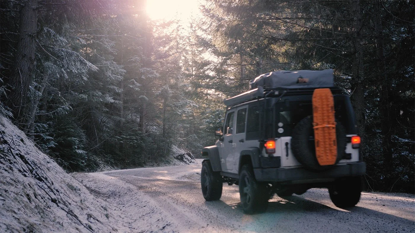 Jeep carrying rooftop gear along a snow-covered forest road, with sunlight filtering through evergreen trees.