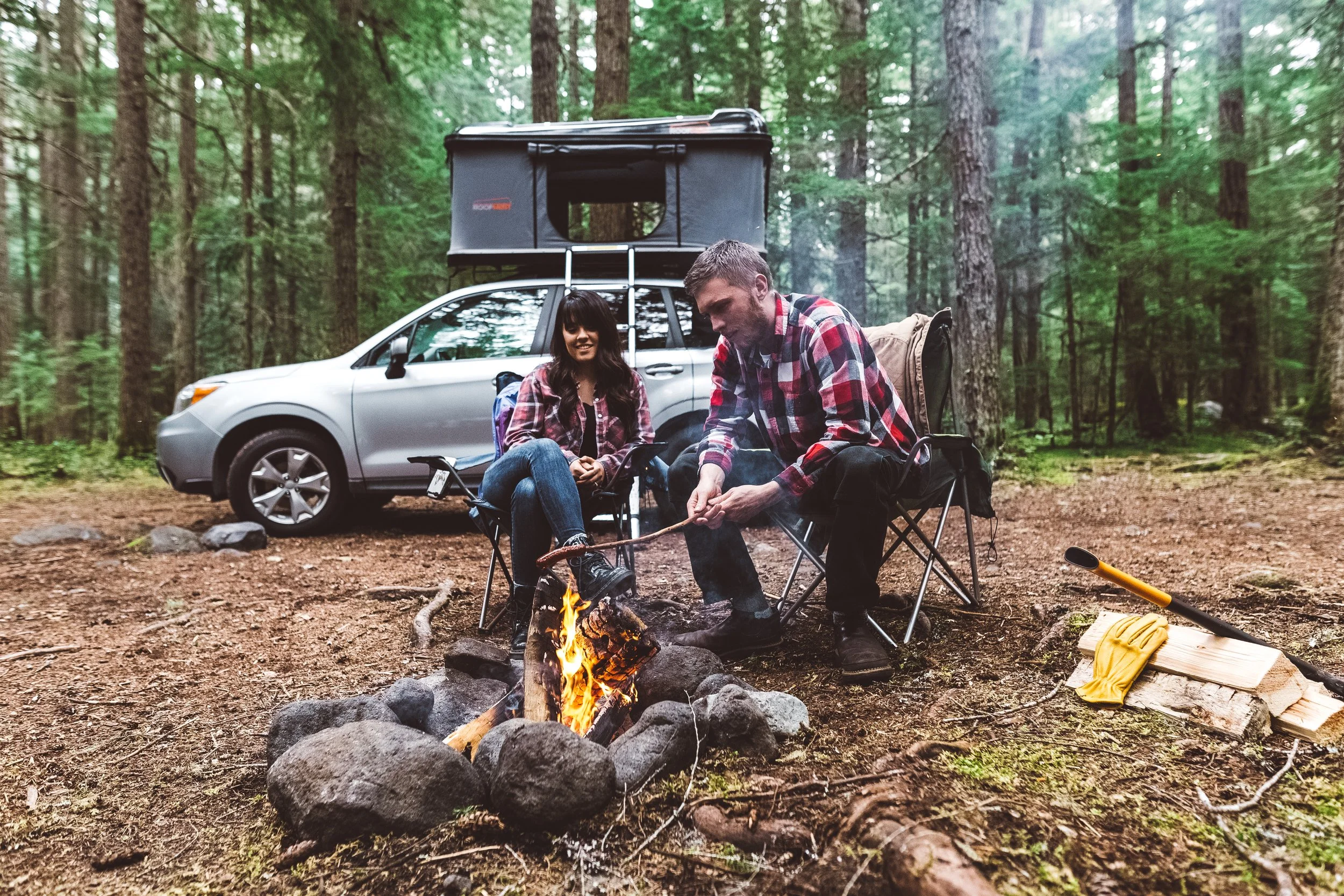 Two people sitting by a campfire in a forest, with a silver car and an over-cab camper on top in the background. They are dressed in plaid shirts and are enjoying the outdoor setting.
