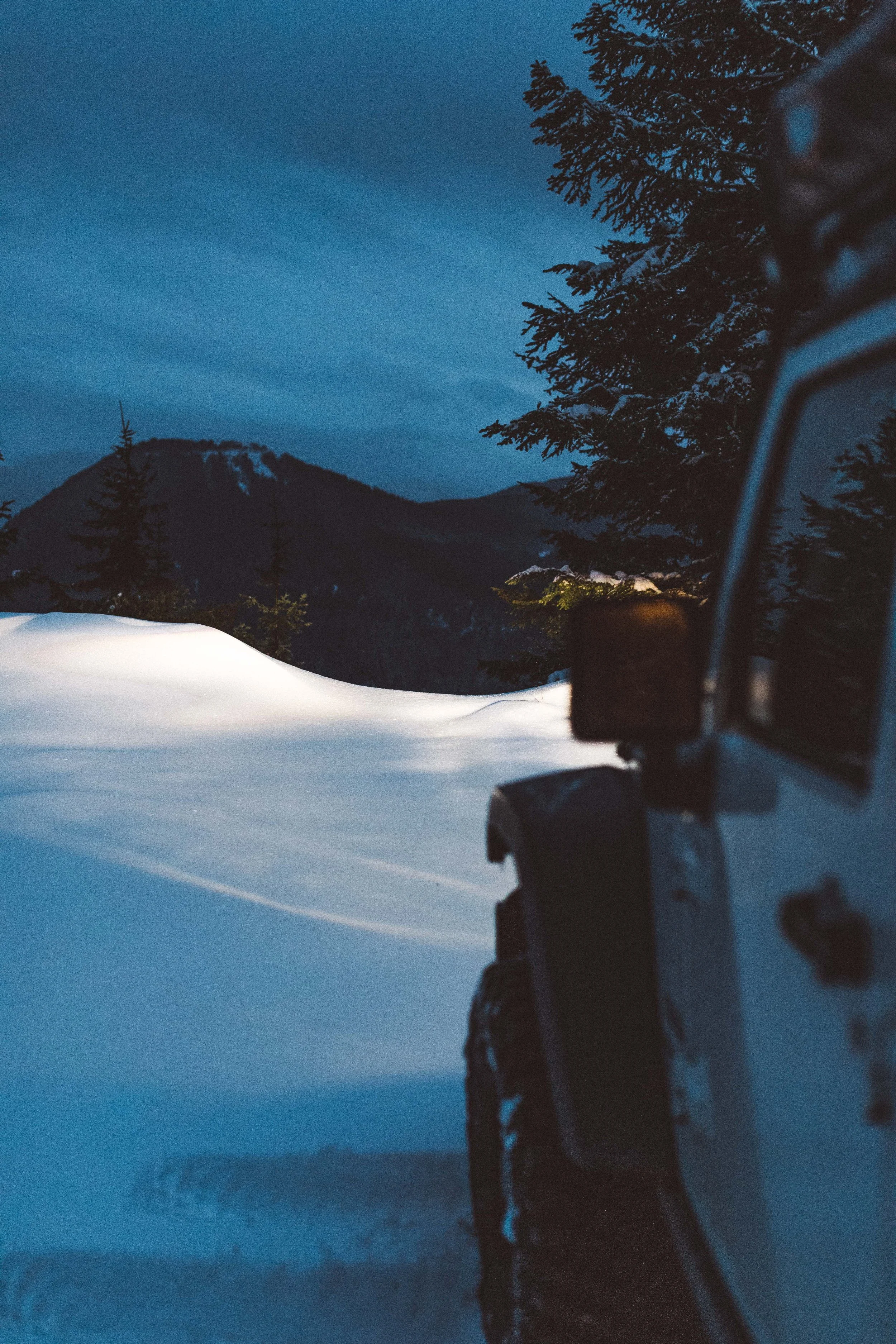 A snow-covered landscape with pine trees and mountains in the background, taken during twilight or early morning, with part of a vehicle visible on the right side of the image.