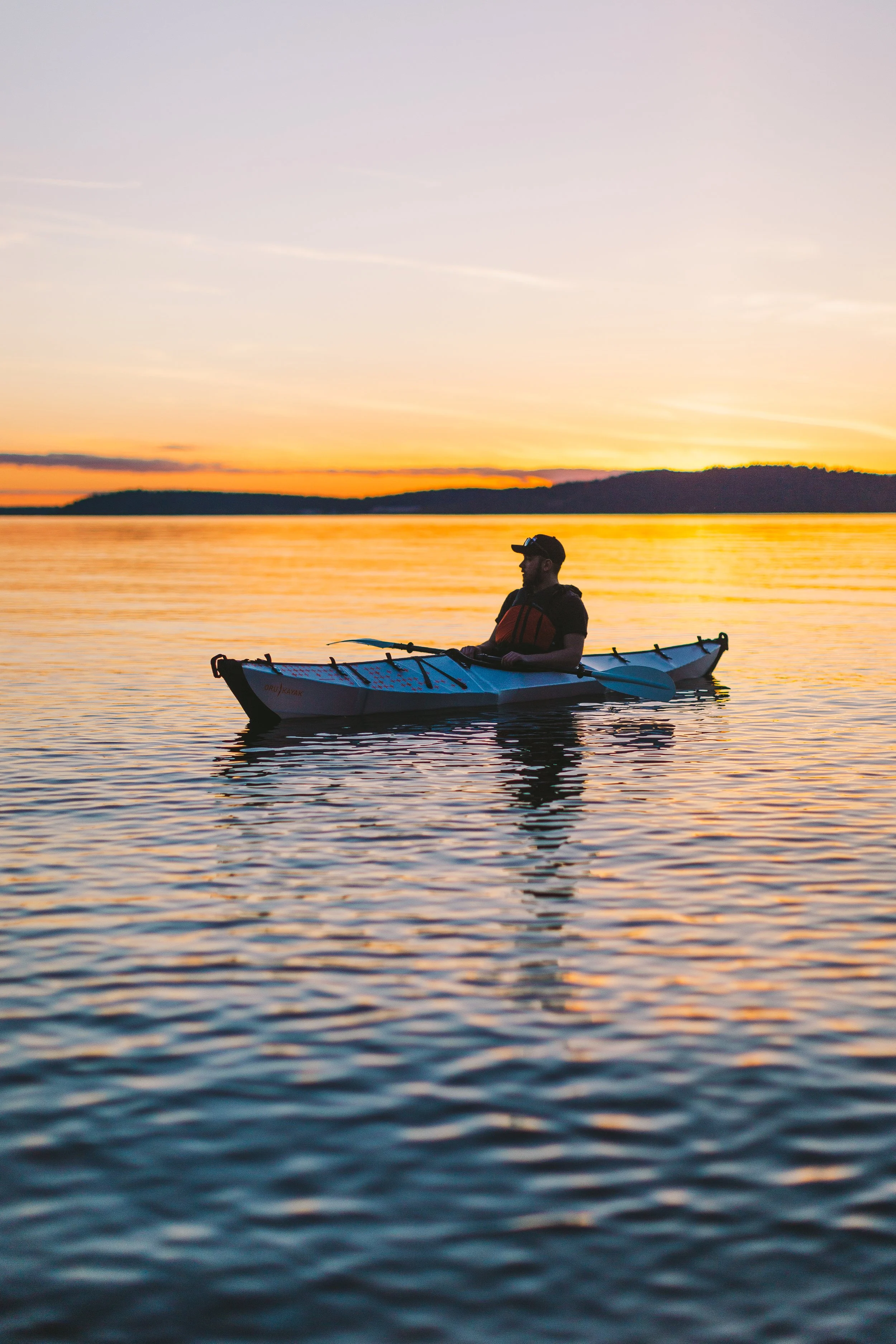 A man kayaking on a calm body of water during sunset with orange and purple skies and a distant shoreline.