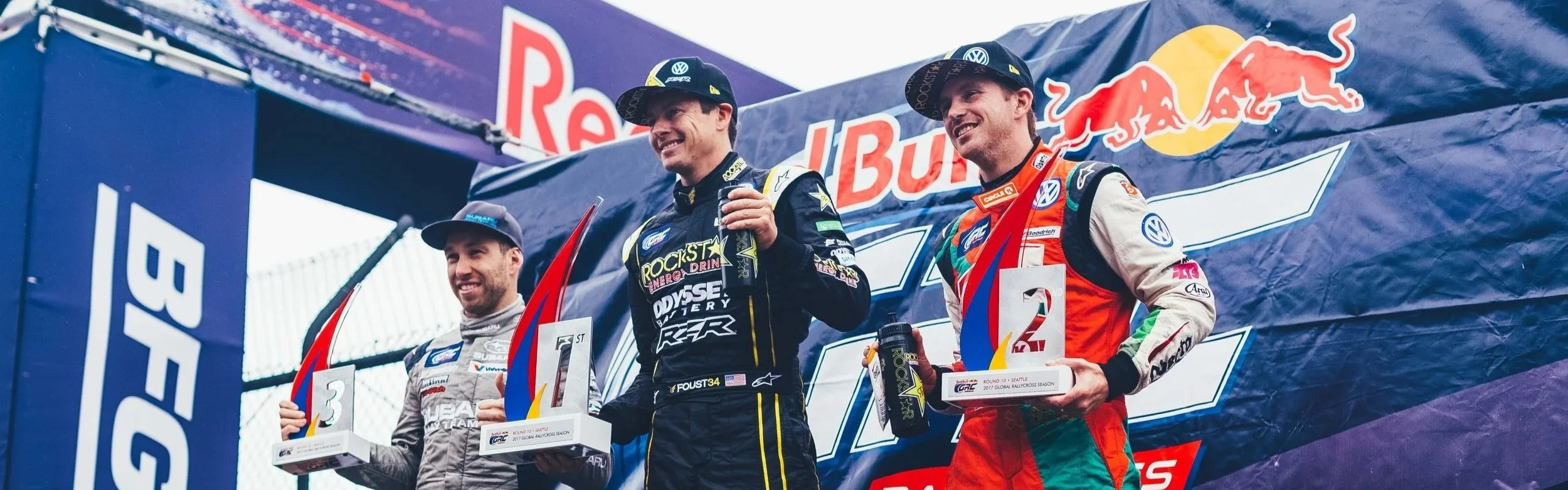 Three race car drivers standing on the podium celebrating with trophies. The driver in the middle, holding a red and blue trophy, is smiling and wearing a black and yellow racing suit. The driver on the right is in a red and white racing suit, holding a trophy, and smiling. The driver on the left is in a gray racing suit, holding a trophy, and smiling. Behind them is a backdrop with Red Bull and other logos.