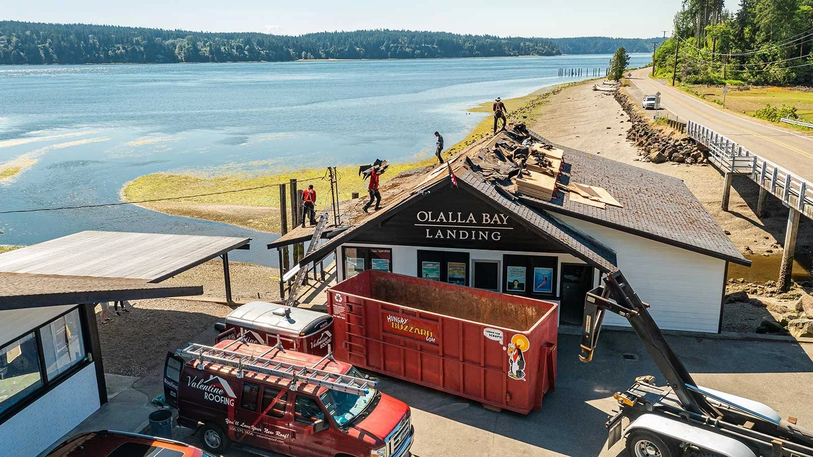 Construction workers repairing the roof of Olalla Bay Landing building near a body of water, with trucks and equipment parked nearby.