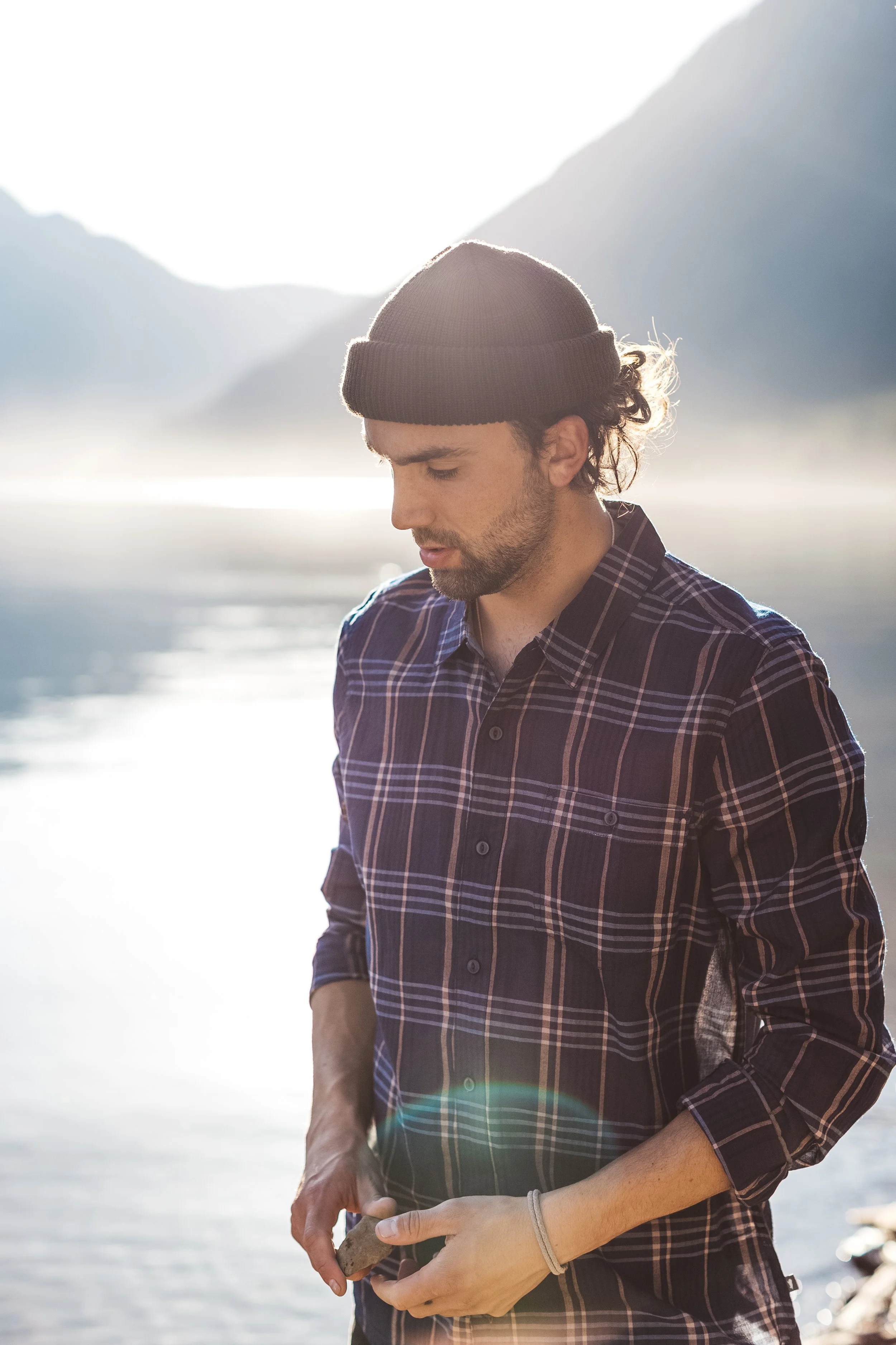 A young man with curly hair, wearing a brown beanie and a plaid shirt, is standing outdoors near water with mountains in the background, holding a small object in his hands.