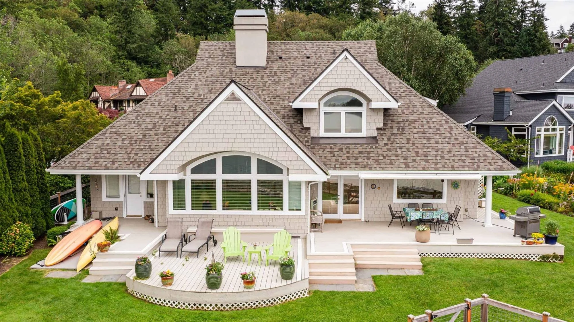 A two-story house with a gabled roof, large front windows, and a patio with outdoor furniture and potted plants, surrounded by lush green trees and neighboring houses.