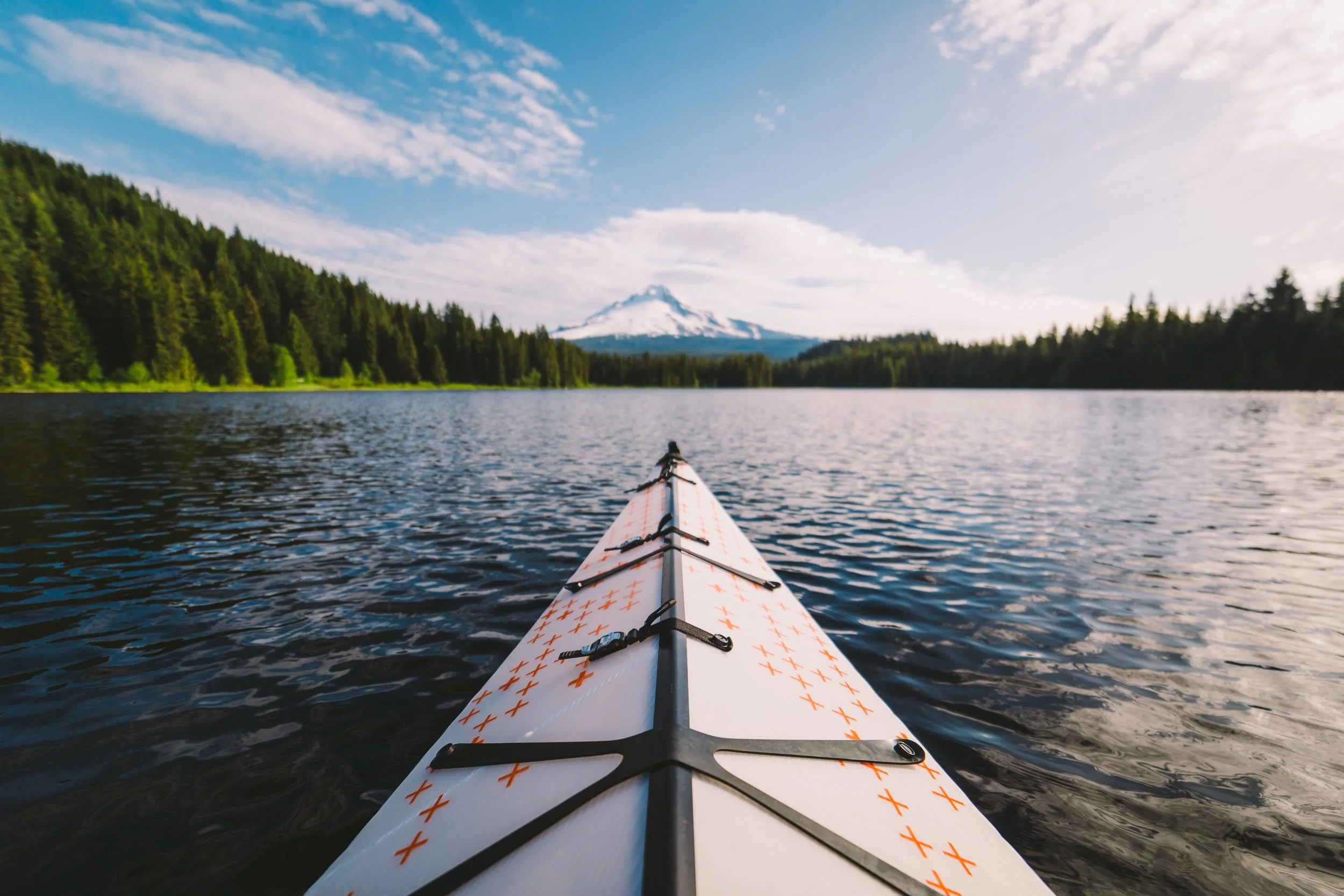 View from a kayak on a lake with a snow-capped mountain in the distance, surrounded by green forest, under a partly cloudy sky.