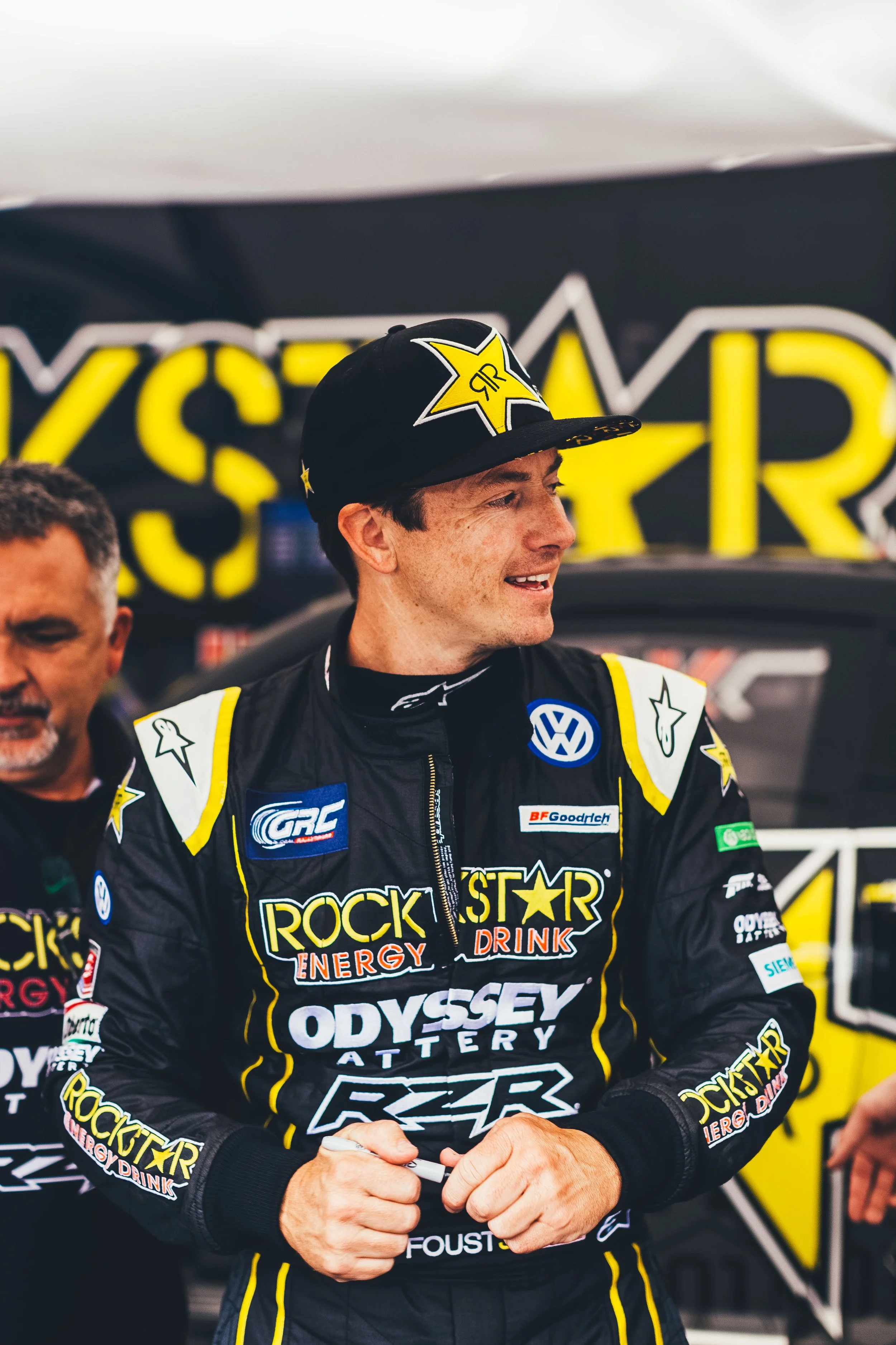 A race car driver in a black racing suit with yellow and white accents, wearing a black cap with a yellow star logo, smiling, standing in front of a yellow and black backdrop with the word 'TEAM' visible.