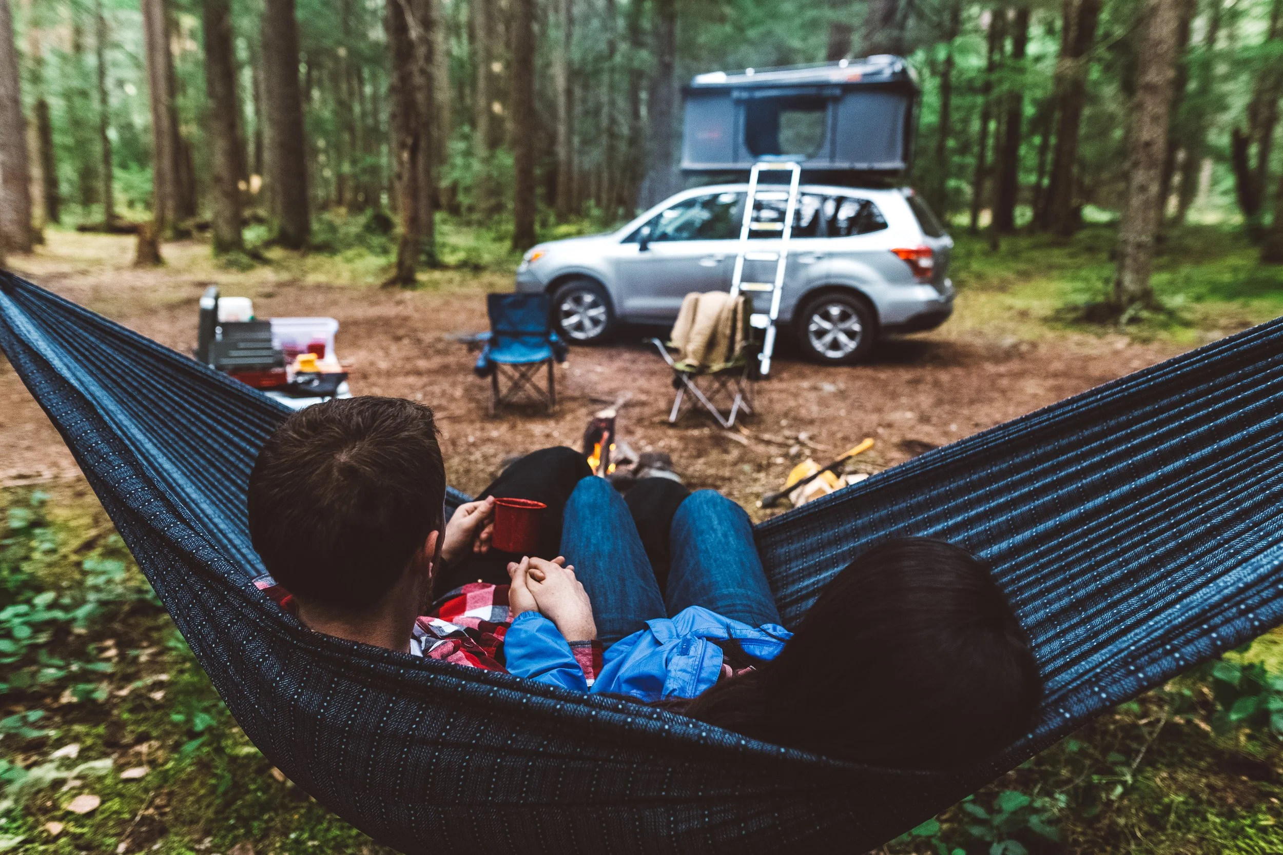 View from a hammock in a wooded campsite showing a person and a child, with a silver SUV and a rooftop tent in the background, along with camping gear and chairs on the ground.