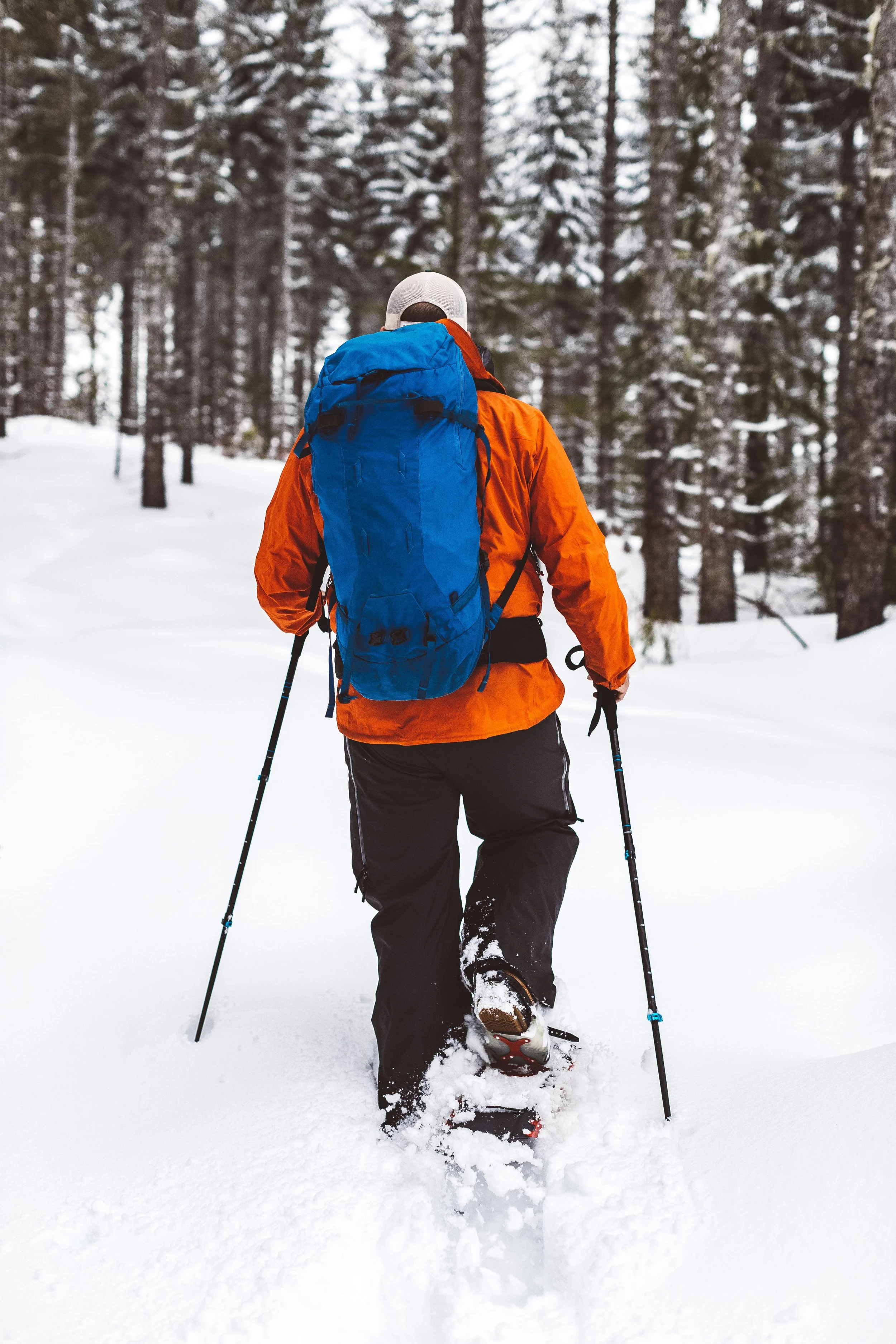 A person snowshoeing through a snow-covered forest in winter, wearing an orange jacket, black pants, a white hat, and carrying a blue backpack.