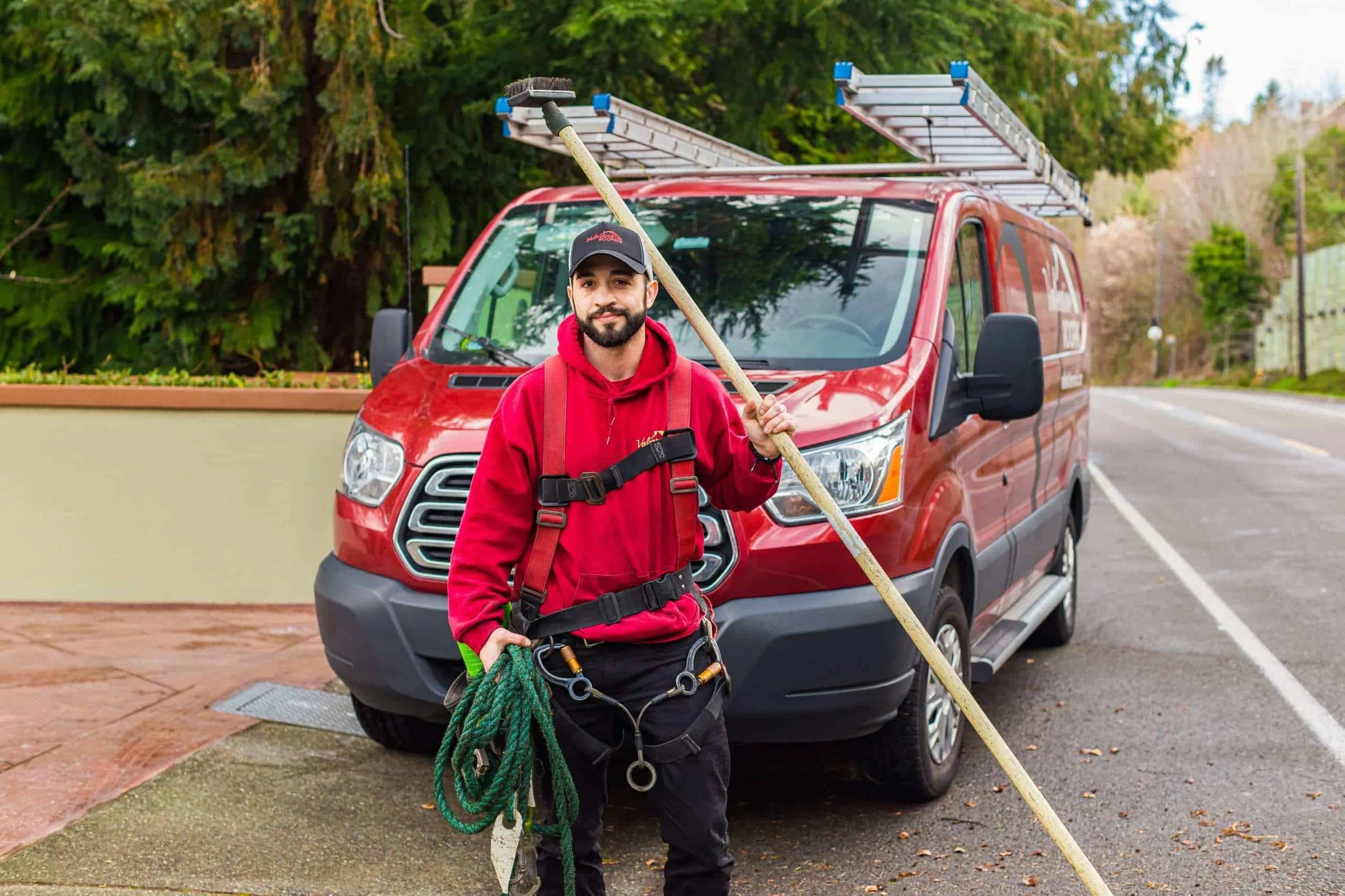 A man in red and black work clothes holding a mop and rope, standing in front of a red van with ladders on top, outdoors with trees and a street in the background.