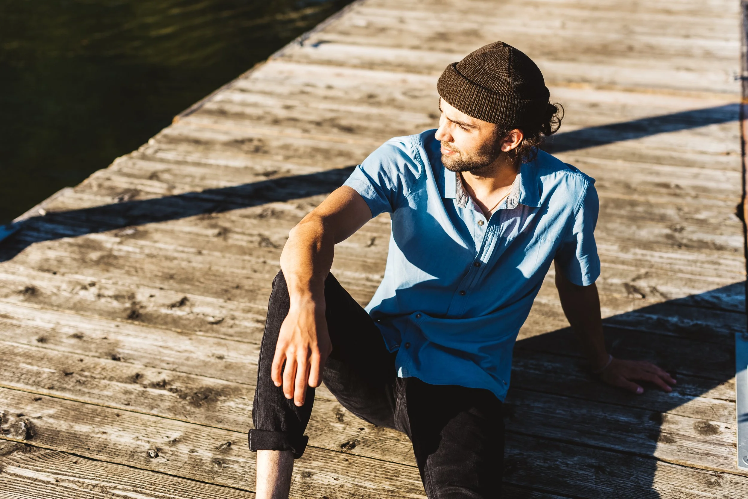 A man sitting on a wooden dock by the water, wearing a brown beanie and a blue shirt, with sunlight casting shadows.