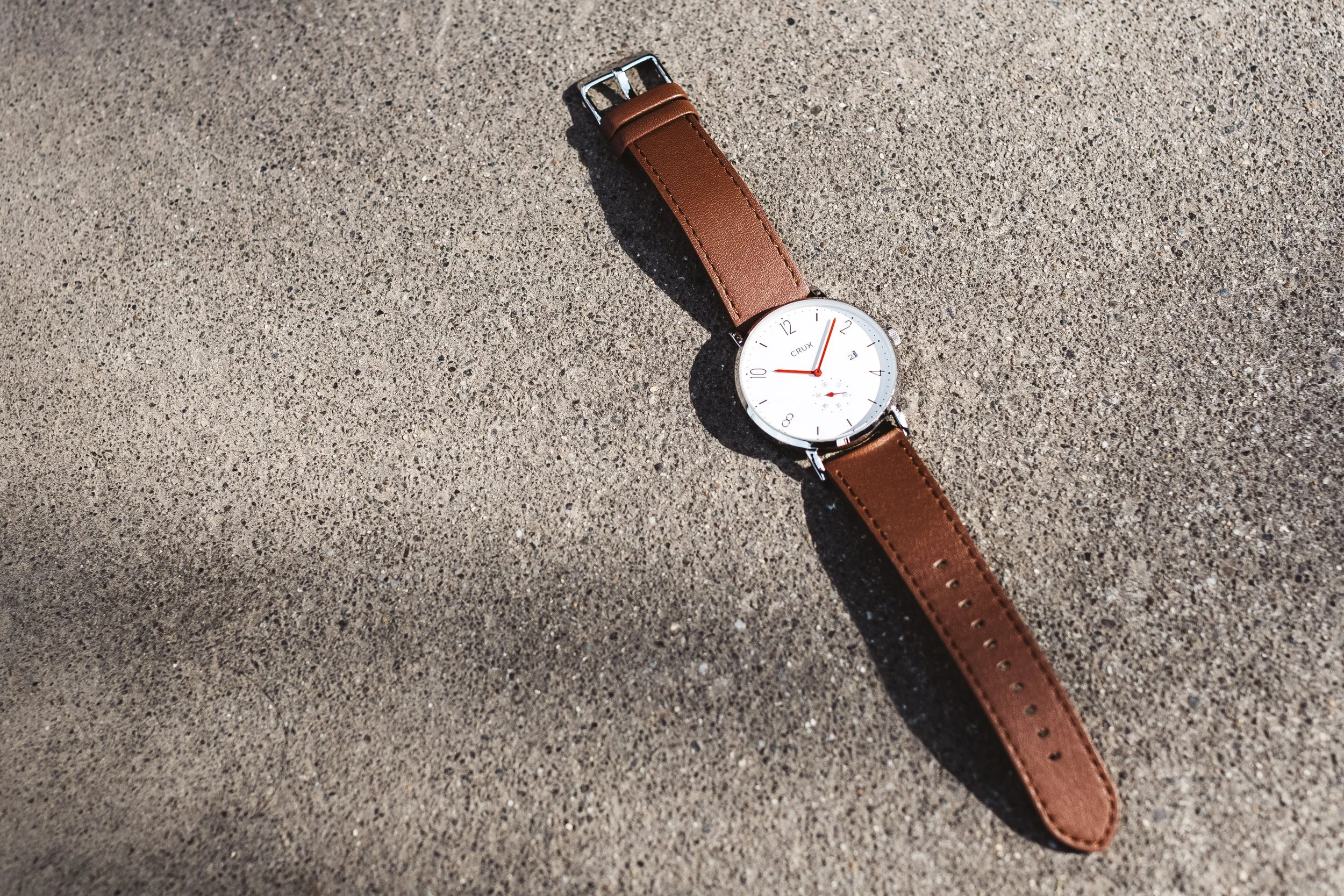 A wristwatch with a white face, brown leather strap, and red second hand lying on textured concrete ground.