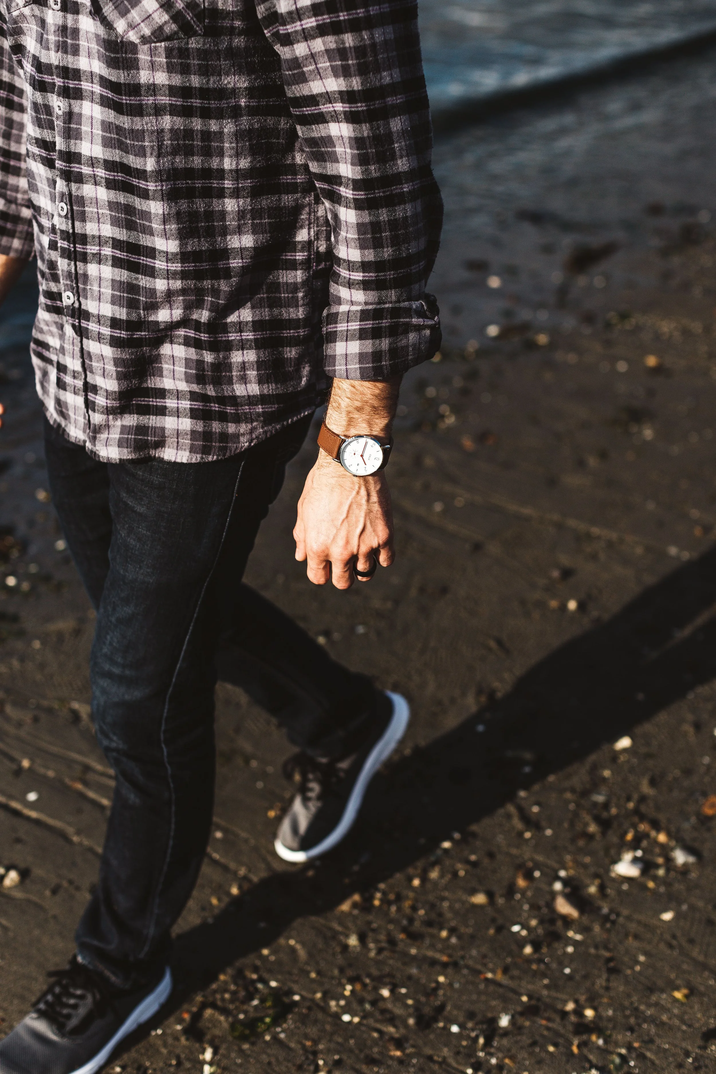 Close-up of a man wearing a casual plaid shirt, dark jeans, sneakers, and a watch on his left wrist, standing on a beach with sand and small rocks.