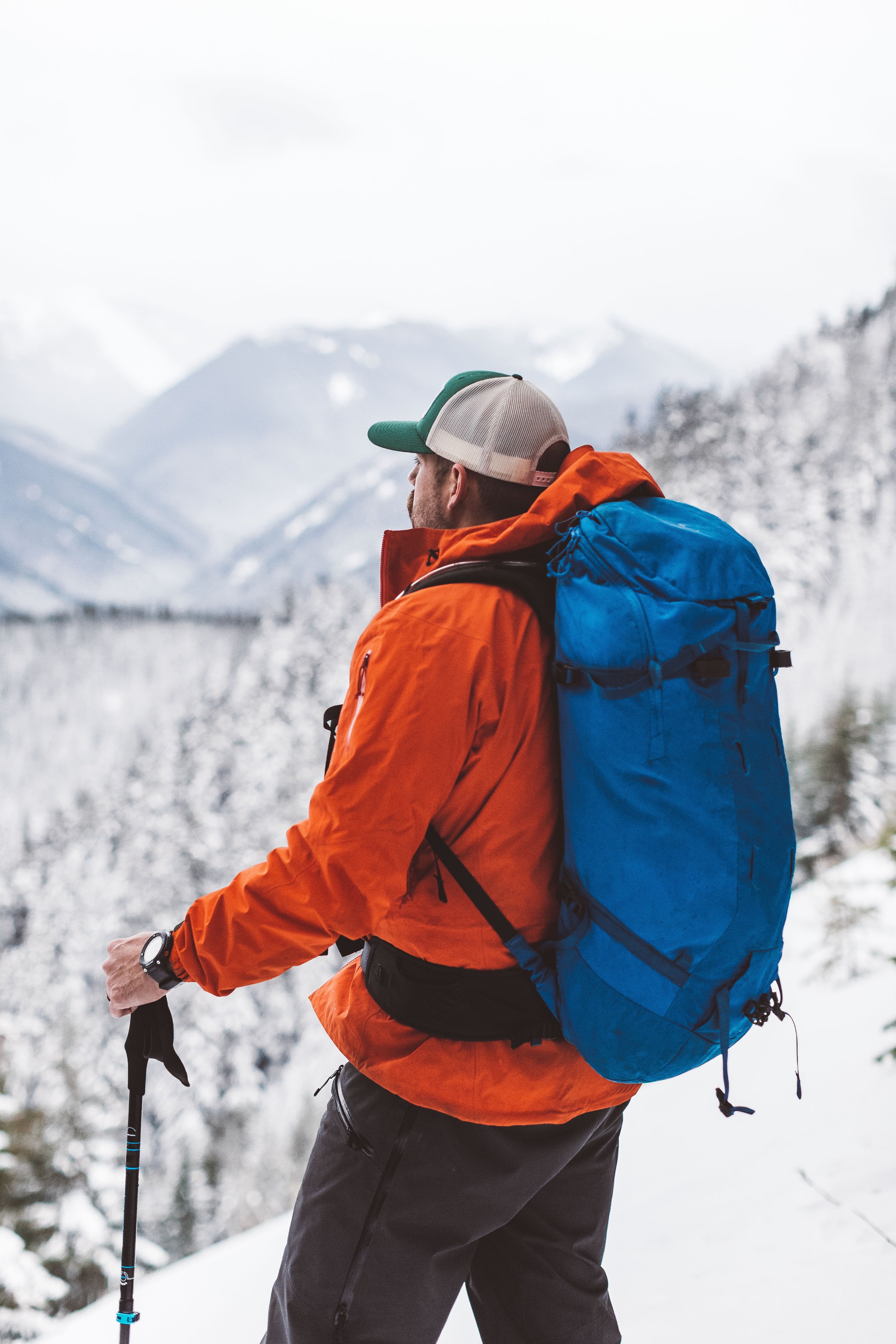 A man dressed for outdoor winter activity, wearing an orange jacket, black pants, a cap, and a backpack, standing in a snow-covered mountainous landscape with snow-capped peaks in the background.