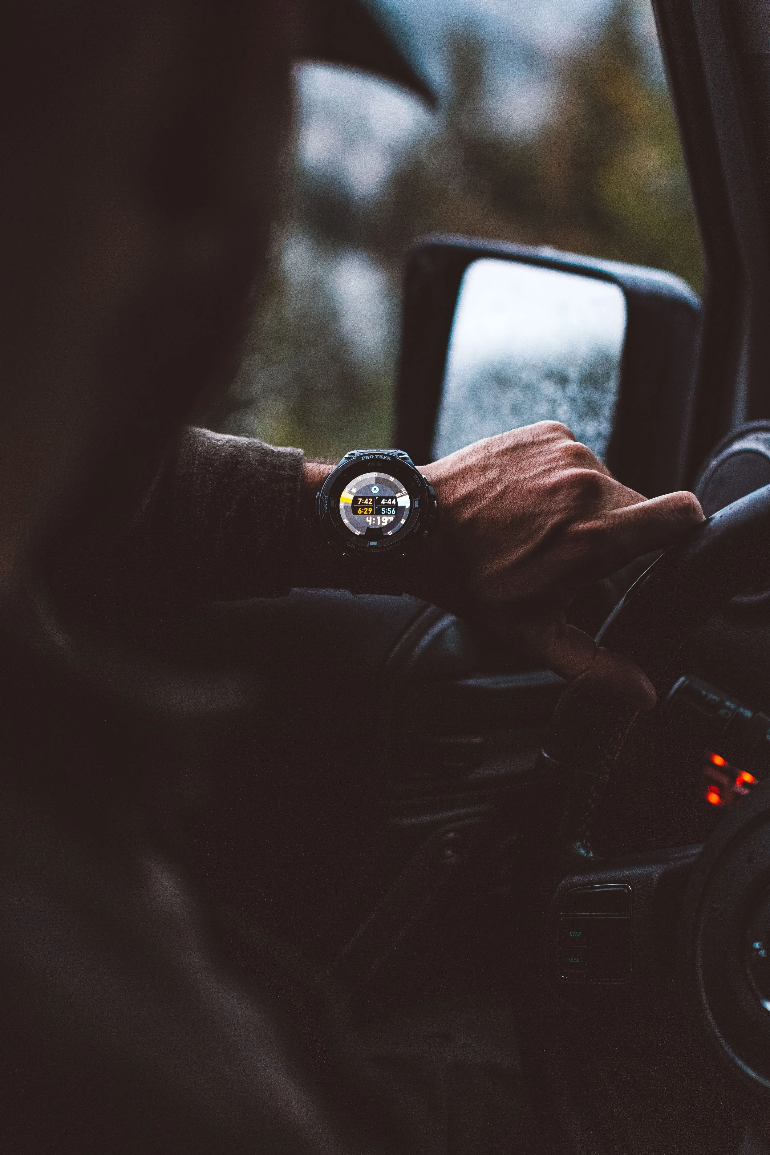 Close-up of a person's hand on a car steering wheel, wearing a black smartwatch displaying multiple data points, inside a vehicle with a blurred view outside the window.