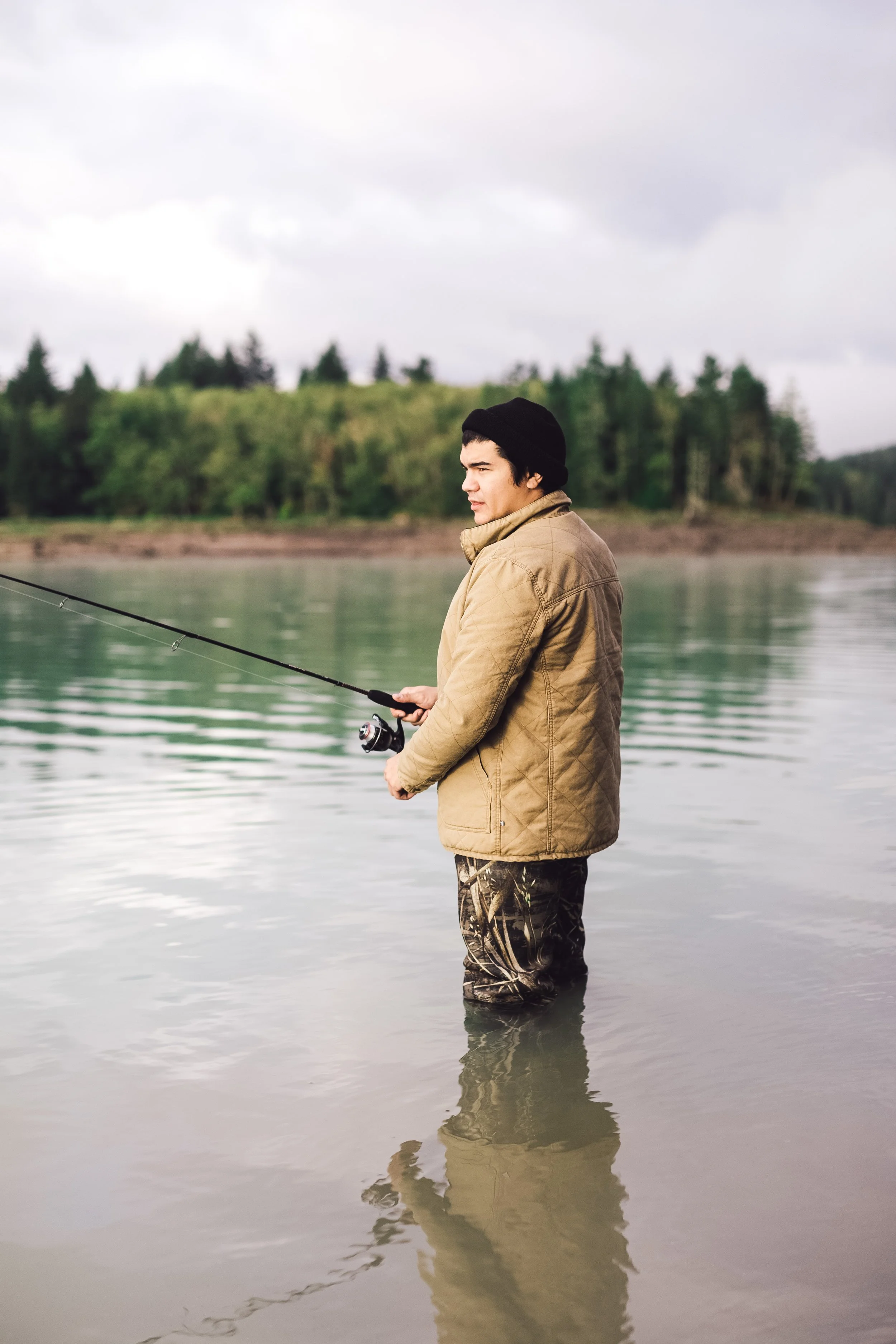 A young man fishing in a river, standing with reflection visible in the water, wearing a black beanie, tan jacket, and camouflage pants, with a background of trees and a cloudy sky.