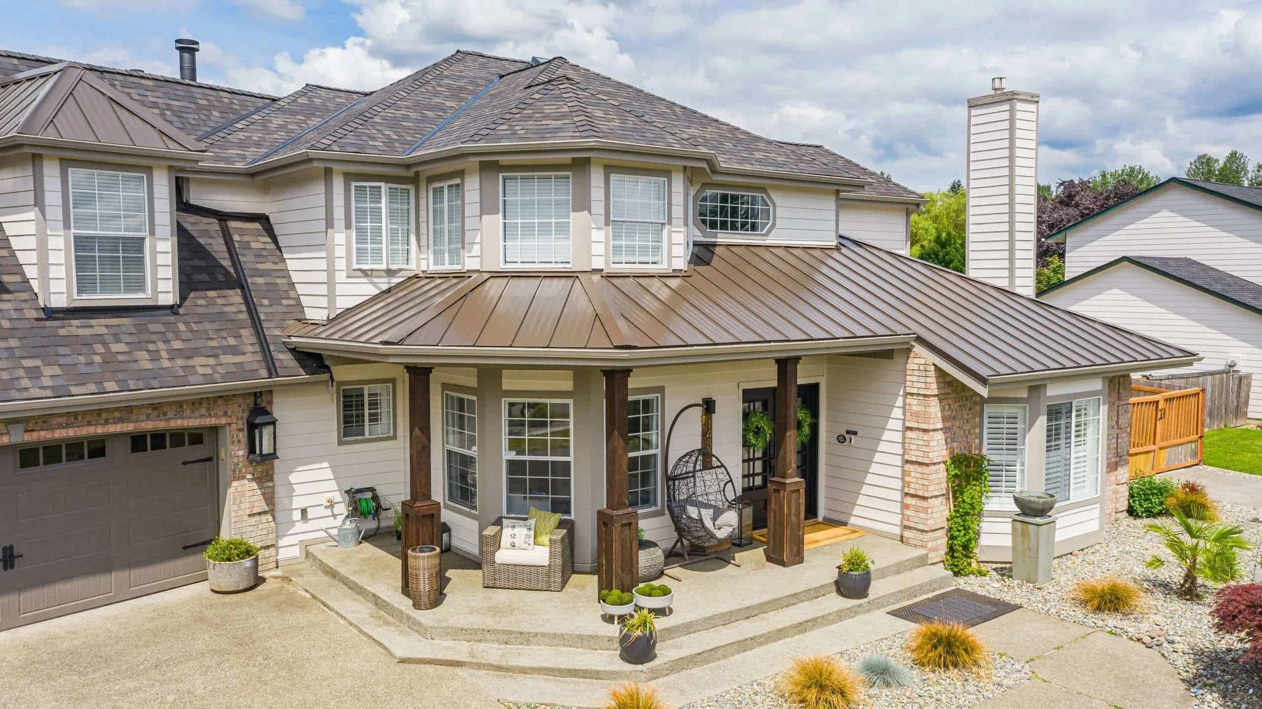 Rear of a house with a brown metal roof, white siding, and a covered patio with outdoor furniture, surrounded by landscaped yard with plants and decorative rocks.