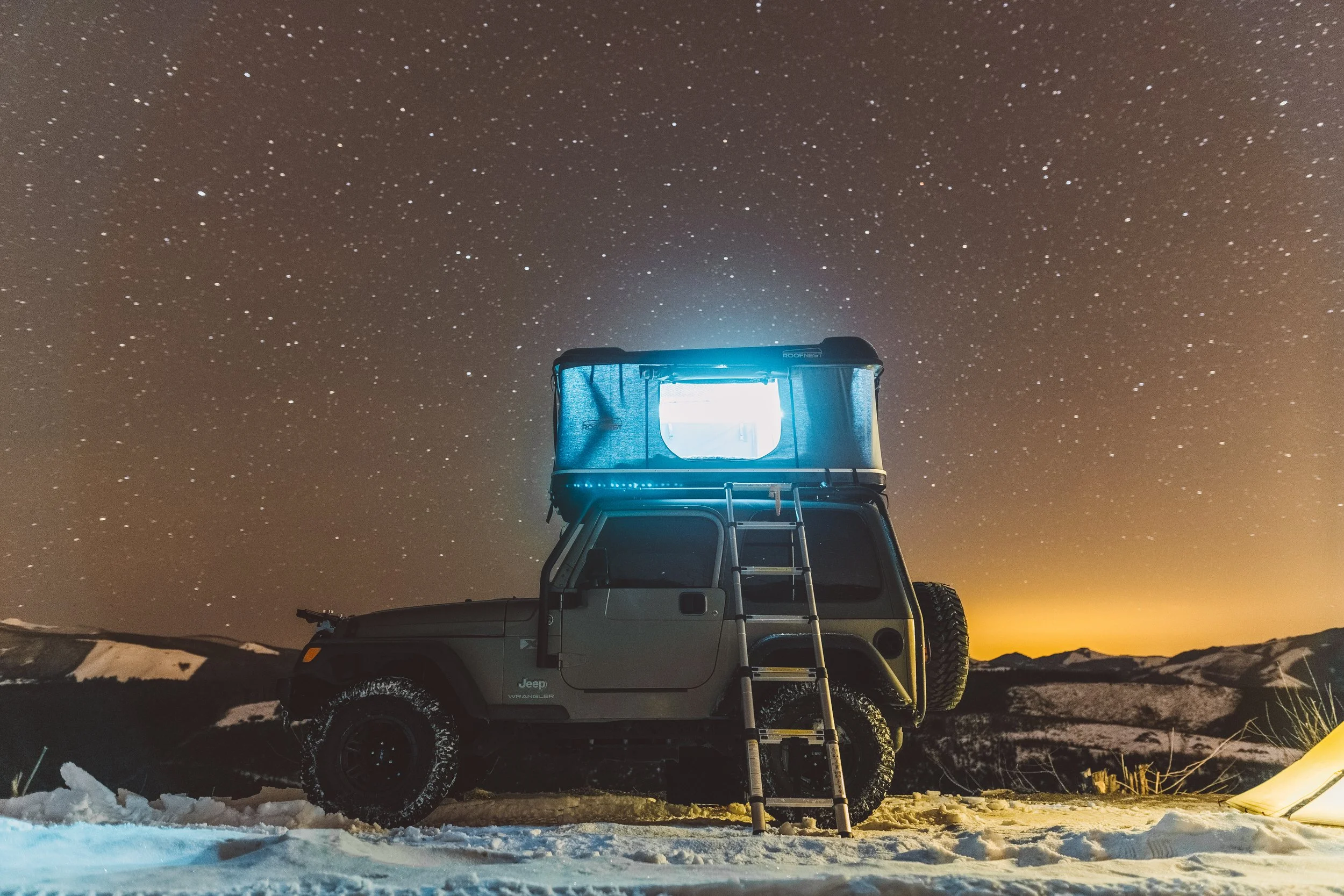 A Jeep Wrangler SUV parked in a snowy landscape with a rooftop tent, illuminated by a bright light, under a star-filled night sky.