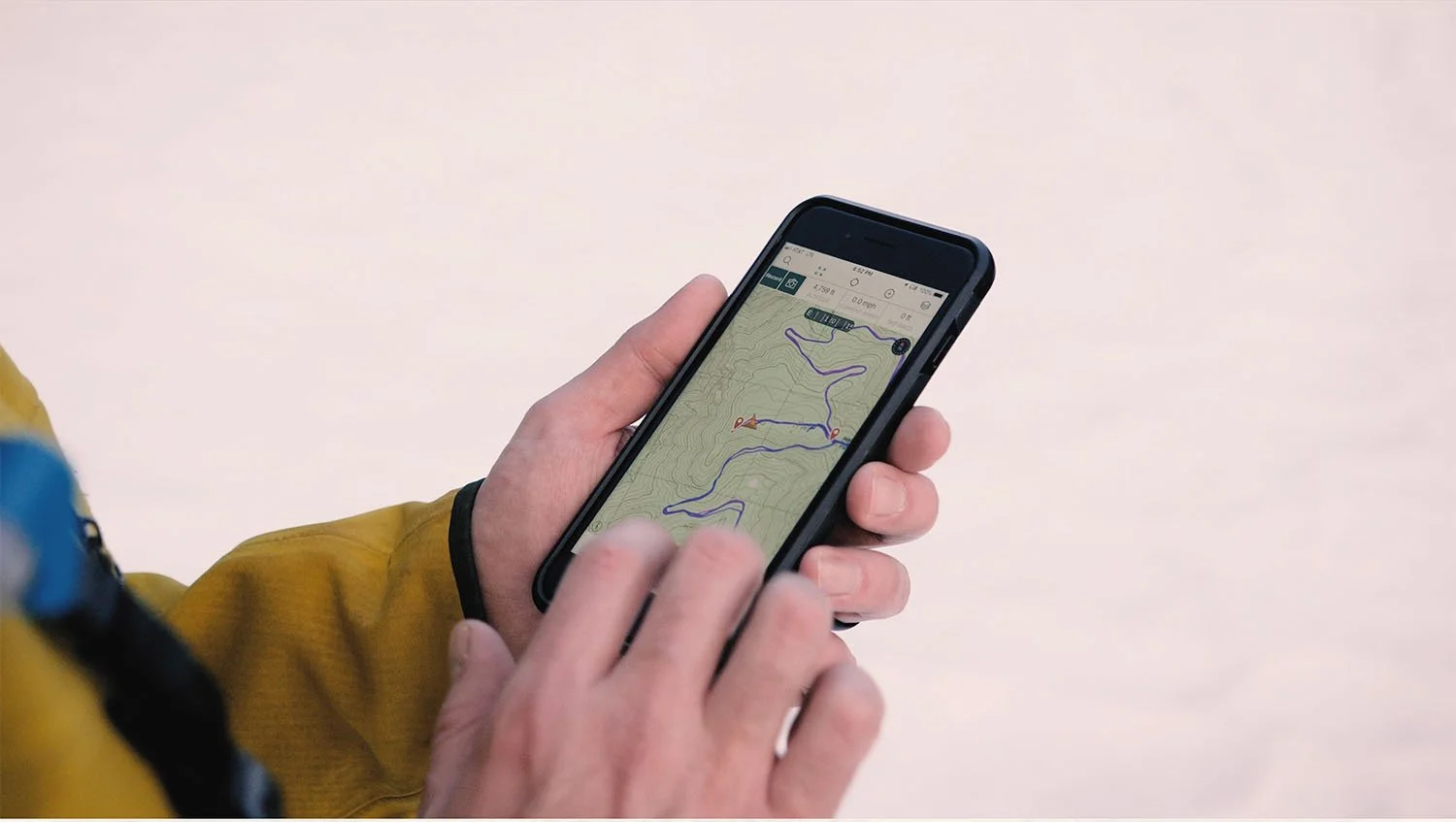 Hiker in a yellow jacket checking a route on a smartphone’s topographic map in snowy conditions.