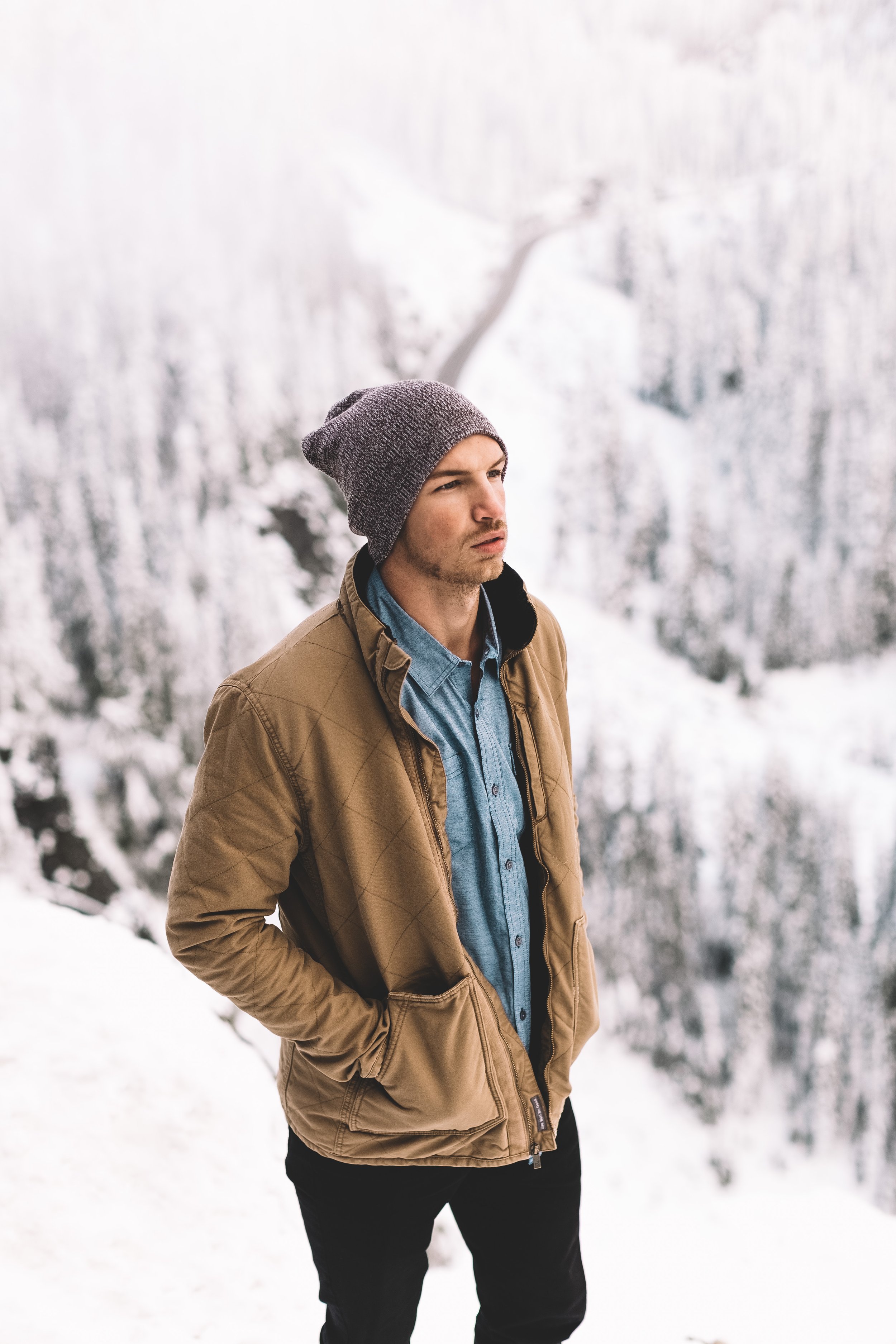 Young man in winter clothing standing outdoors in a snowy landscape with snow-covered trees in the background.