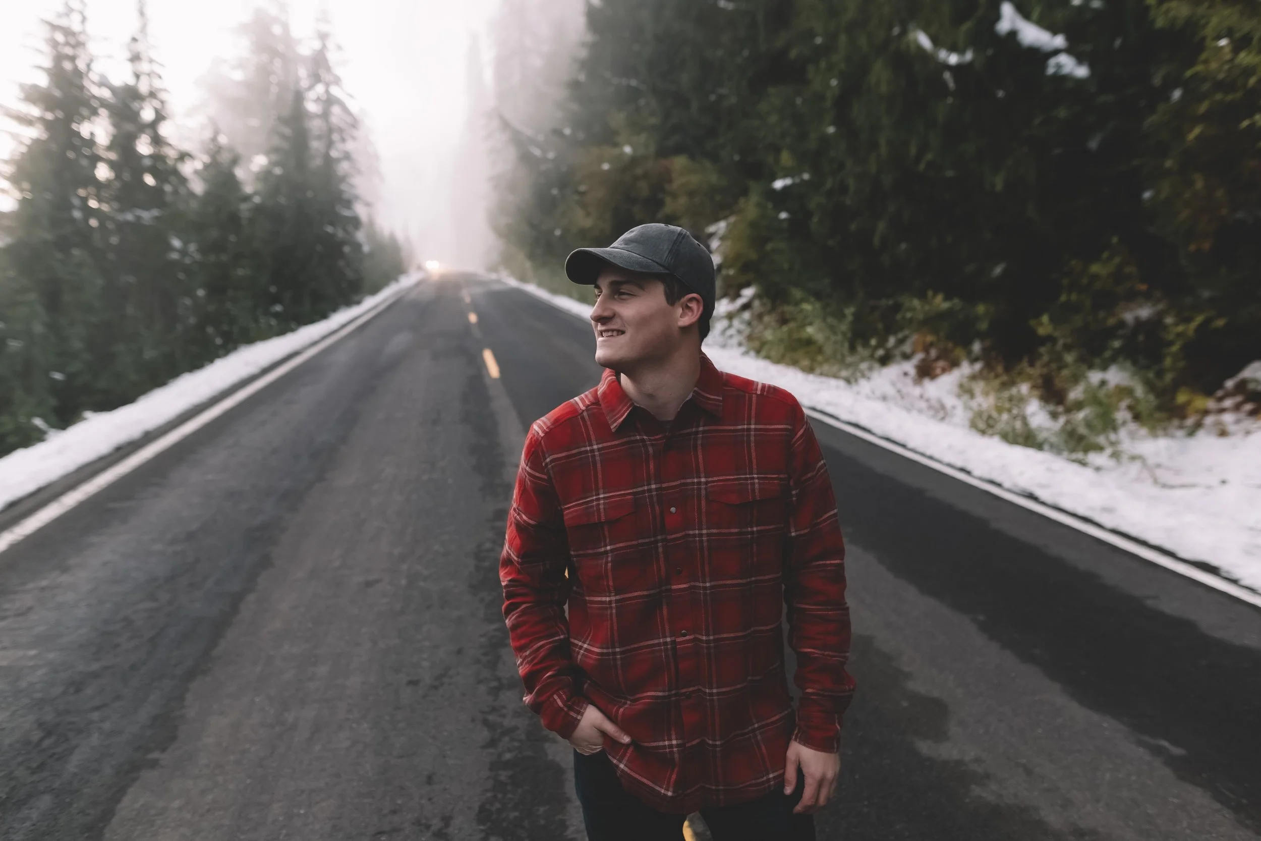 A young man wearing a grey cap and red plaid shirt walking on a foggy mountain road with snow on the sides and dense trees in the background.