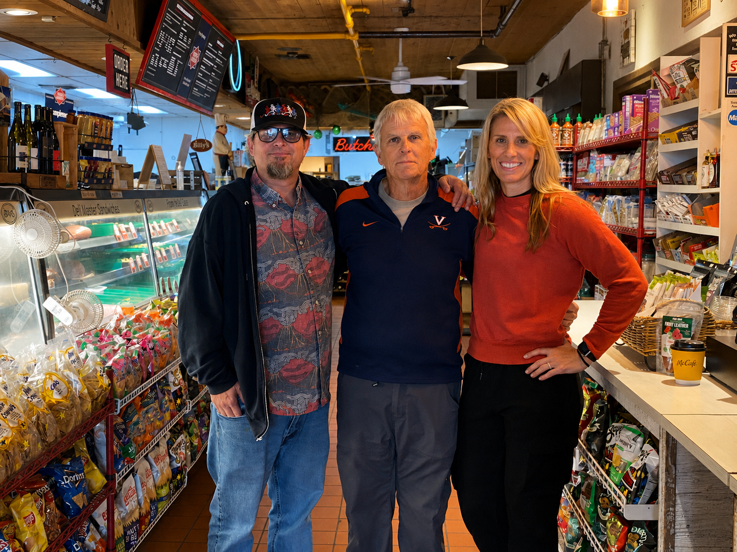 Three people pose inside a deli and convenience store surrounded by snacks, drinks, and grocery shelves.