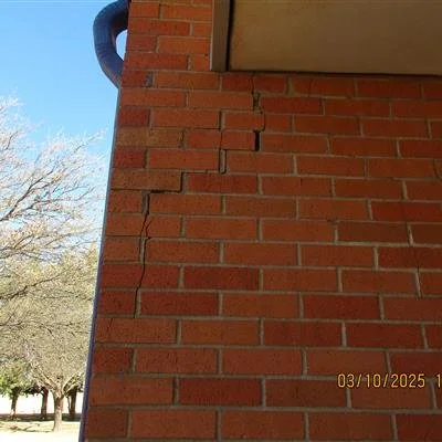 Close-up of a brick wall with visible crack on the corner of a building. Part of the building's overhang and a tree with bare branches are visible in the background.