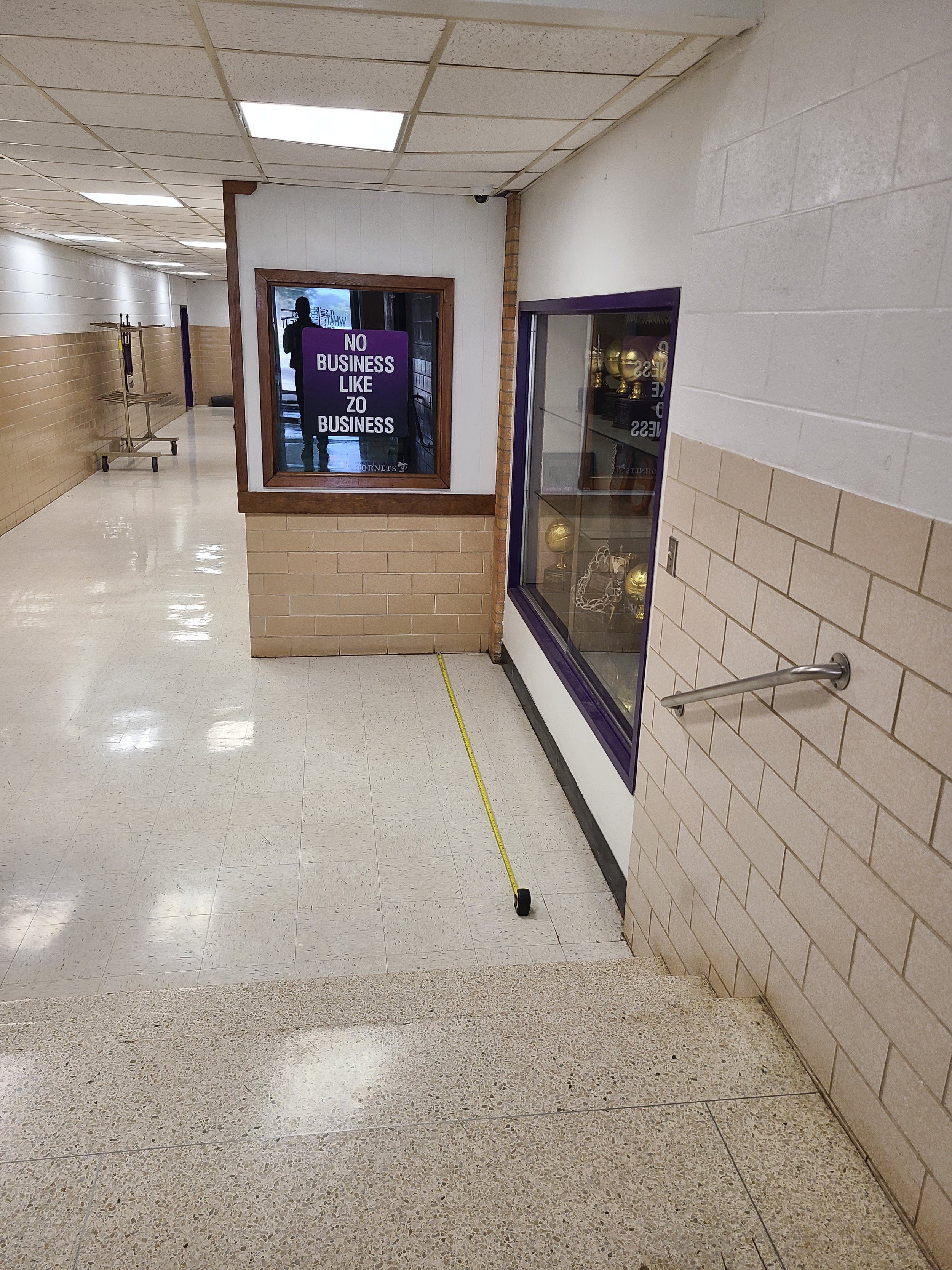 Empty school hallway with beige and white tiled walls, a coat rack, a window display case with sports trophies, and a yellow measuring tape on the floor.