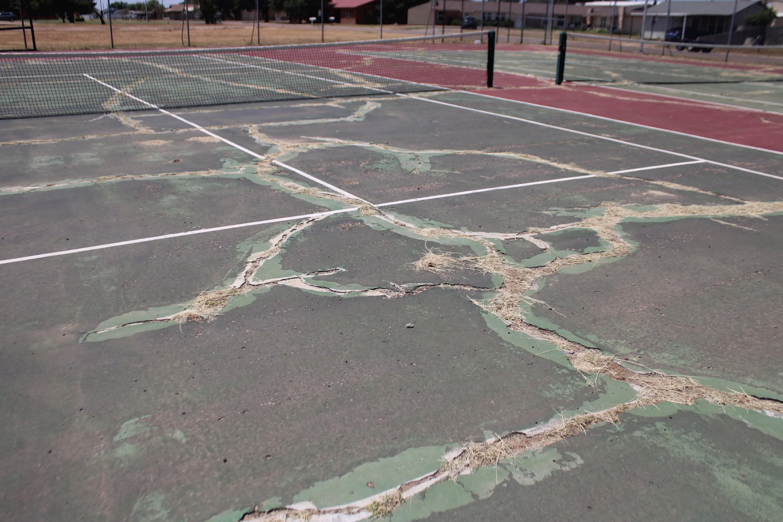 A damaged outdoor tennis court with faded and cracked surface, torn net, and overgrown weeds in the court cracks.