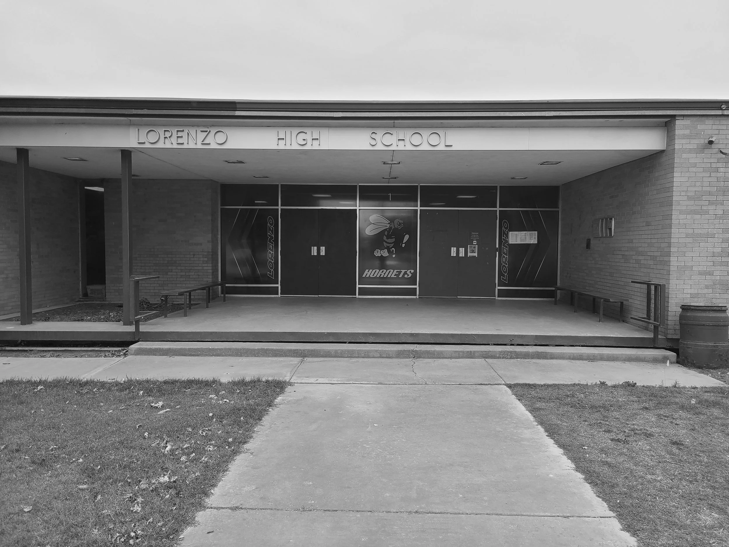 The entrance to Lorenzo High School, a brick building with large glass doors and a bench outside. The school's name is displayed above the entrance.