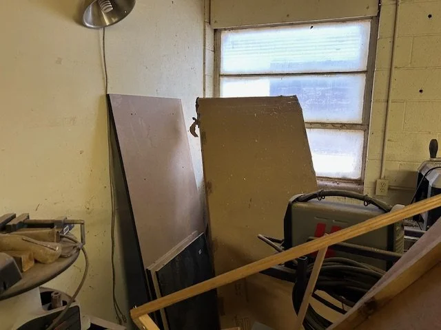 A cluttered workshop corner with a large frosted window, a tilted brown door, and various tools and materials, including a lamp, a handheld power tool, and wooden planks.