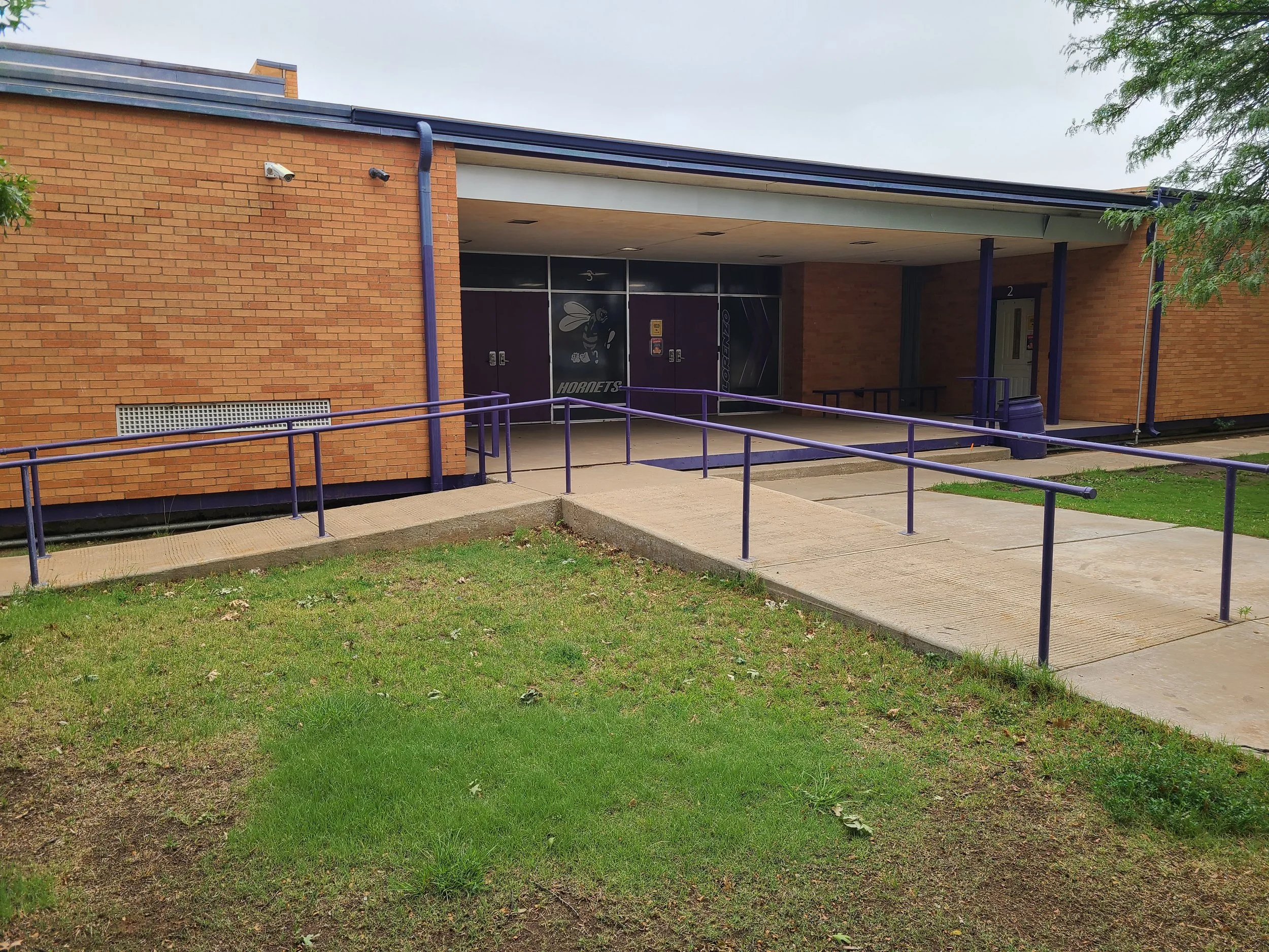 Exterior of a brick building with purple railings, windows, and a door, with grass and concrete in the foreground.