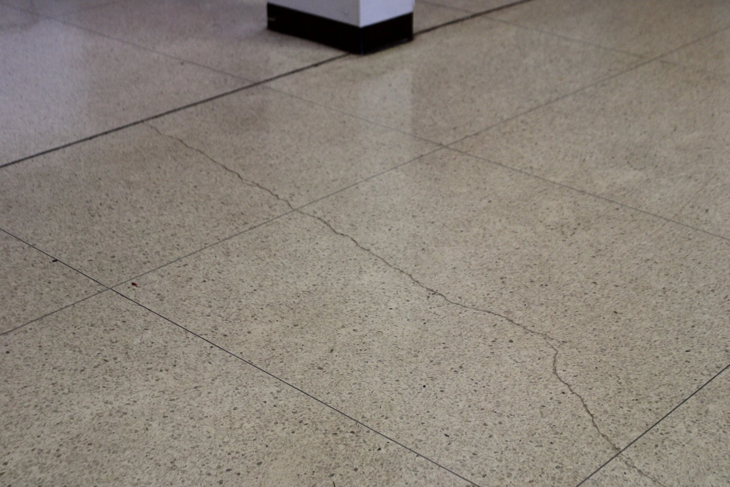 Close-up of a speckled beige tile floor with visible cracks and dark grout lines, and a corner of a piece of furniture in the upper part of the image.
