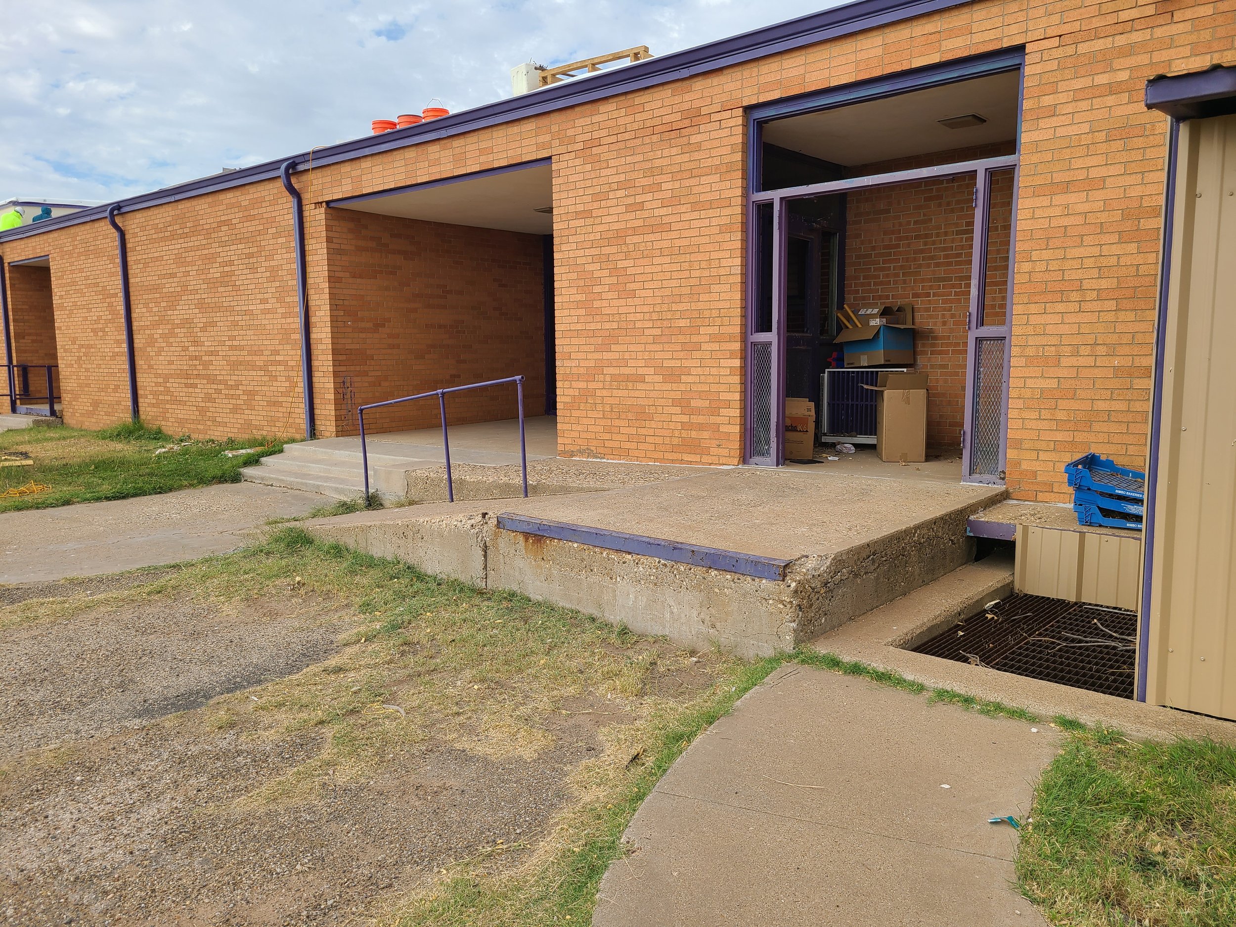 Exterior of a brick building with purple window frames and a small concrete steps leading to a doorway. There is construction or storage materials inside the building visible through the open door. The sky is partly cloudy.