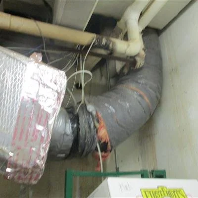 A damaged large black duct pipe with insulation material exposed and broken in an industrial or mechanical room.
