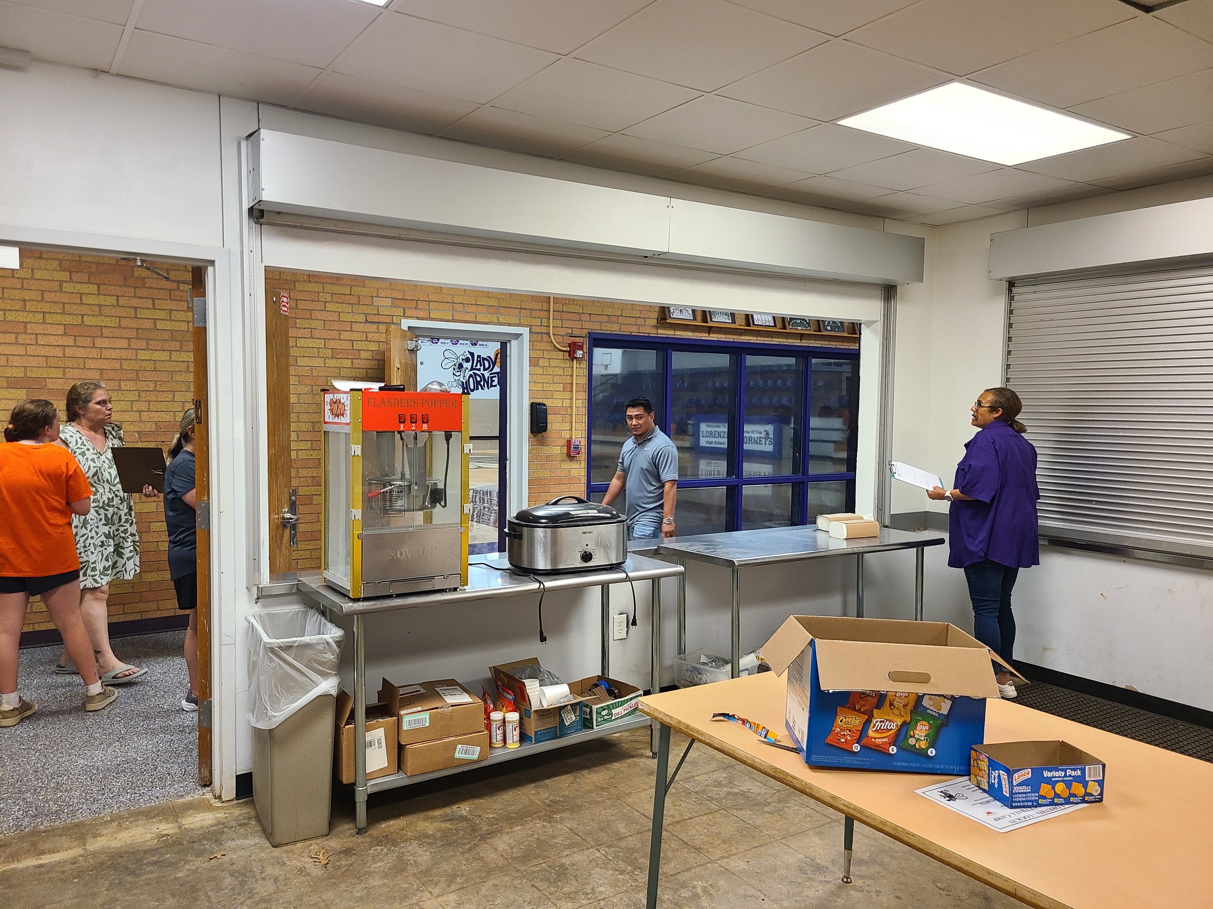 People standing inside a room near tables with various supplies, snack chips in boxes on one table, a popcorn machine on a metal table, and boxes underneath, with a brick wall and glass door in the background.