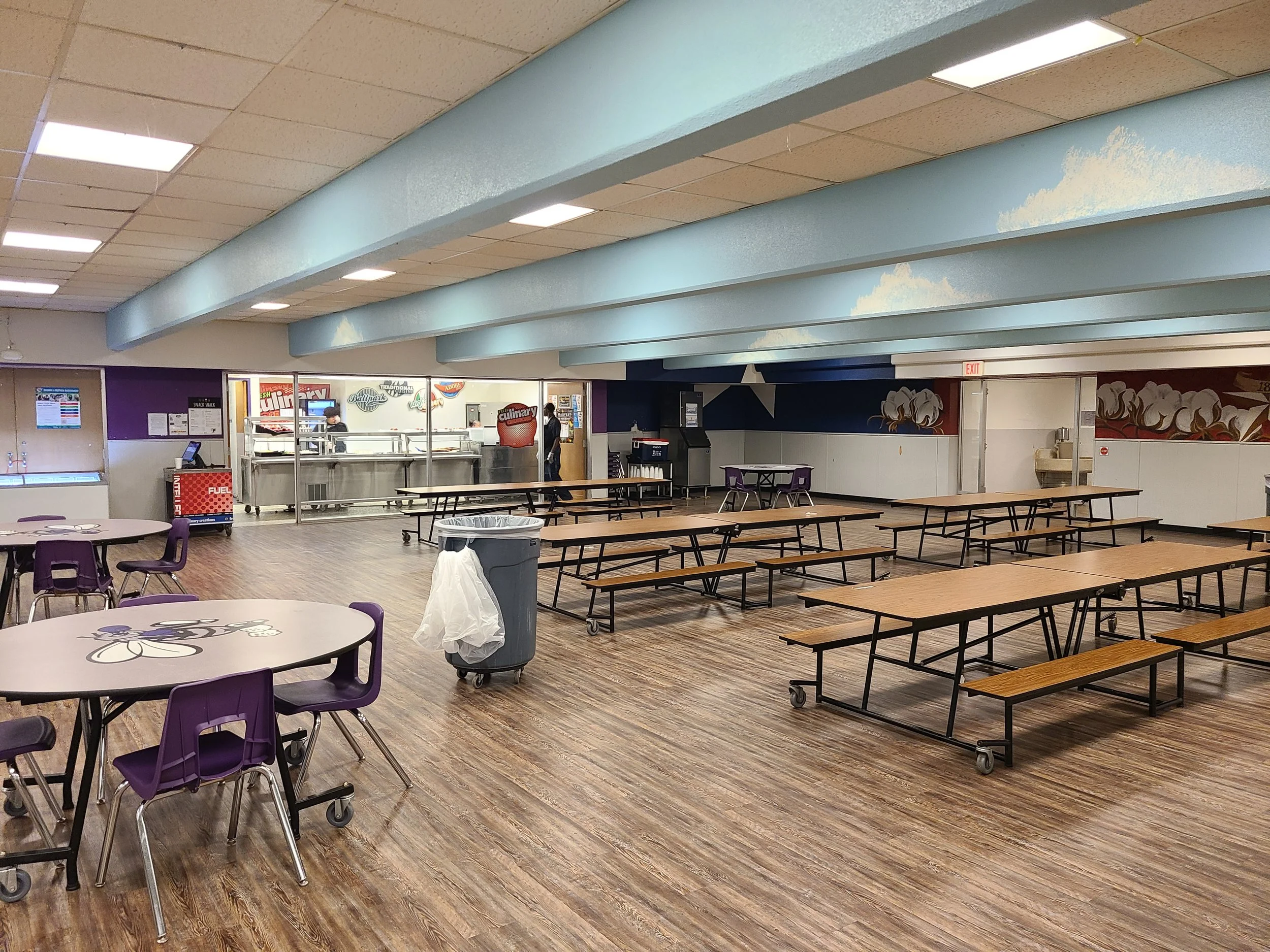 Empty cafeteria with wooden floors, purple chairs, and long wooden tables. A trash can and a few people in the background near a food service area with a mural and signage.
