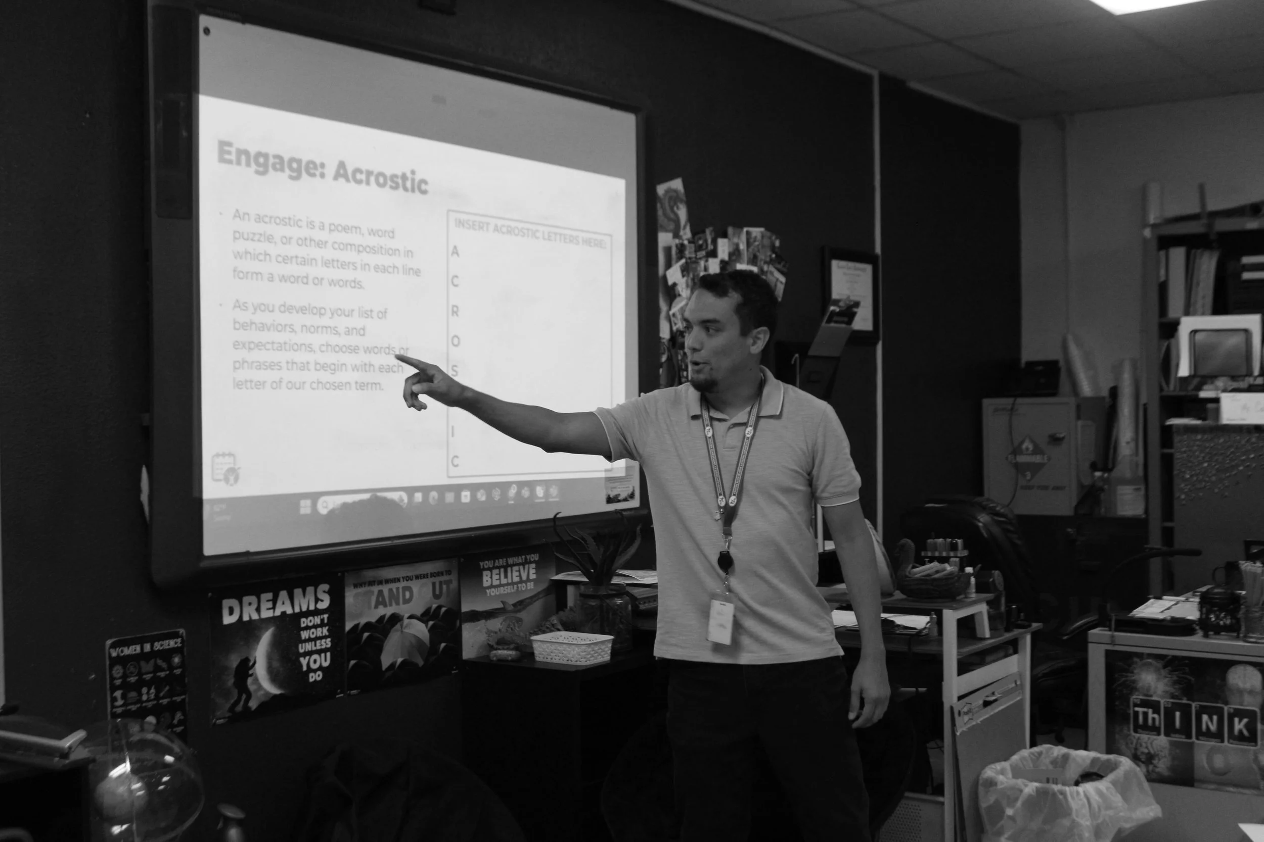A man is standing in front of a screen giving a presentation about acrostic poems in a classroom or office setting. There are posters, shelves, and office supplies visible in the background.