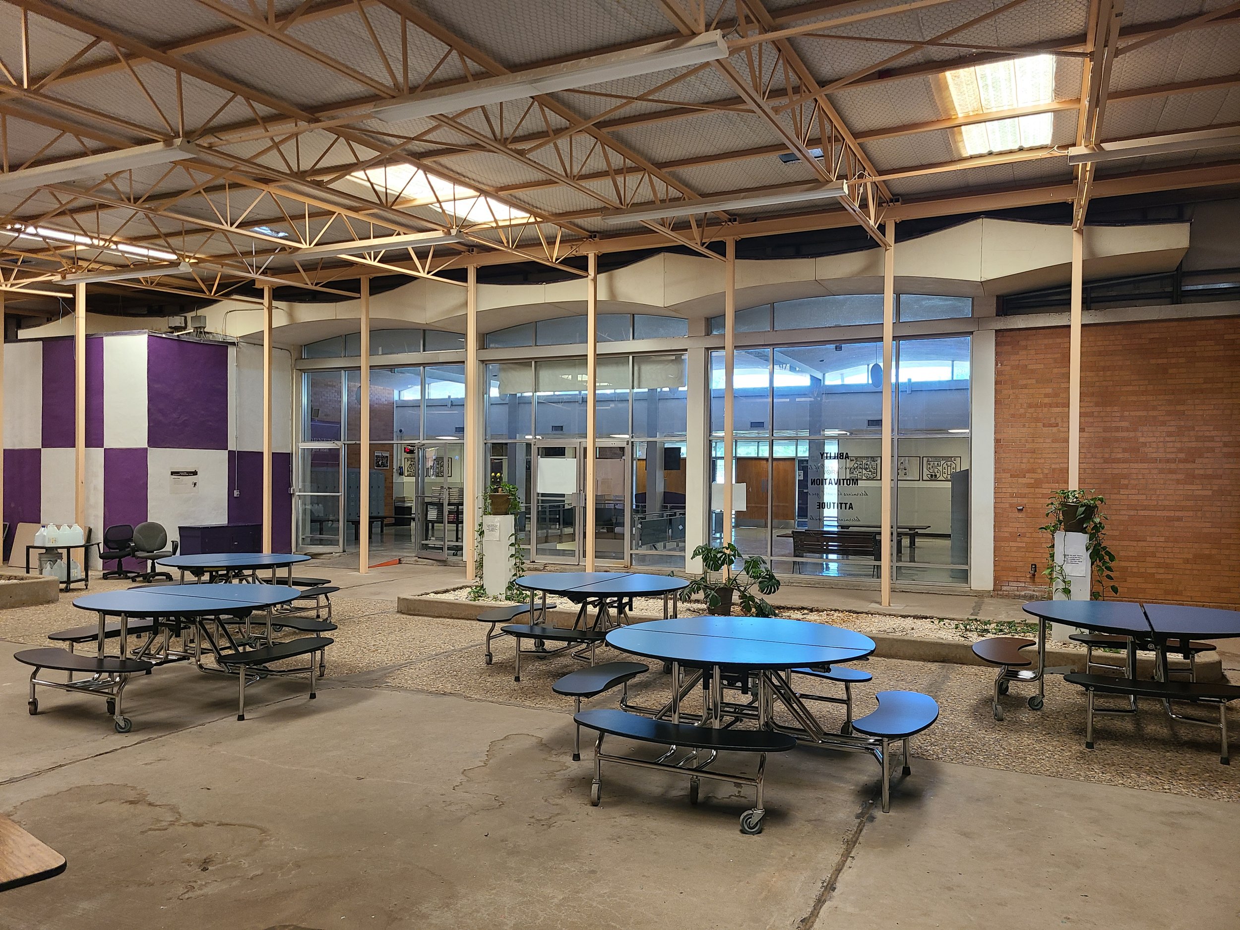 Indoor space with folding tables and chairs, brick and glass walls, and a ceiling with exposed lattice structure and skylights.