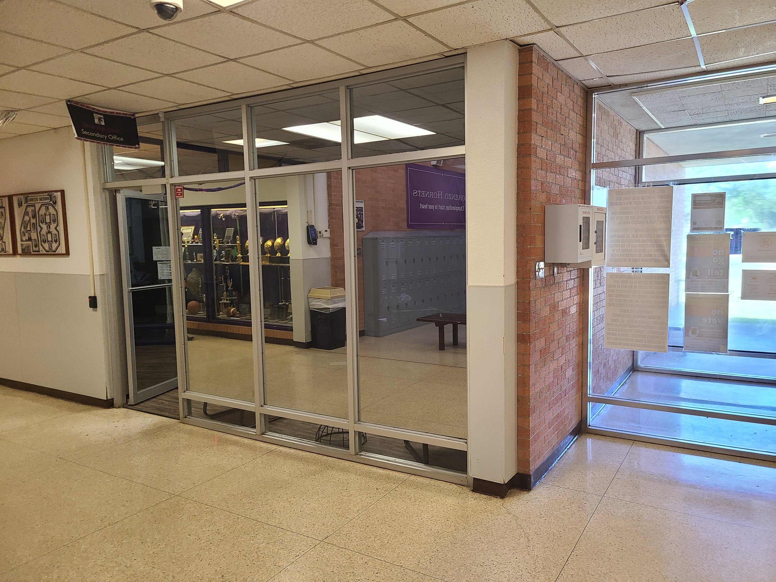 Entrance of a school or office building with glass doors, a brick wall, and signs indicating 'Secondary Office' and other notices inside.