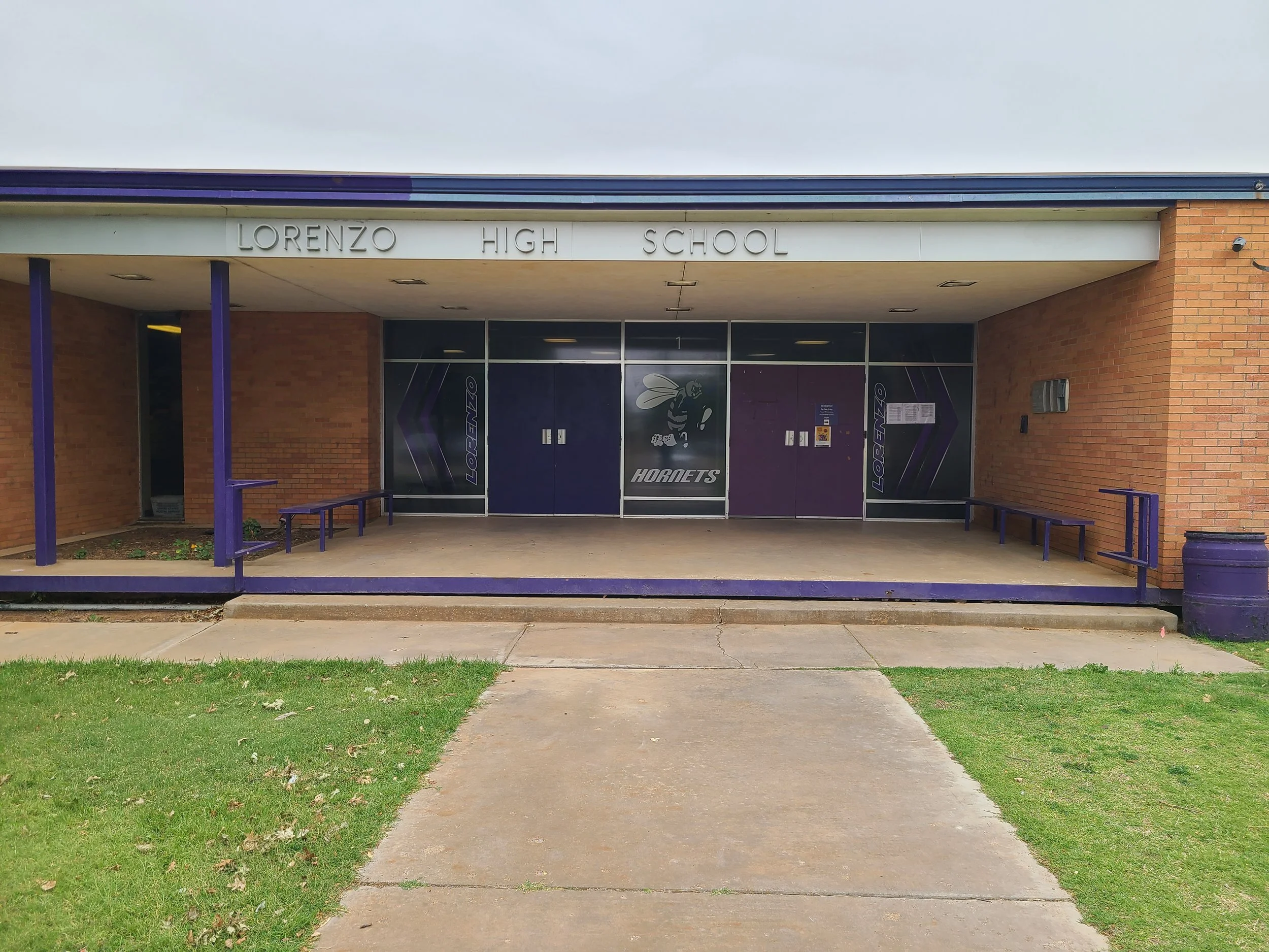 Front entrance of Lorenzo High School with purple benches and a purple bee mascot on the glass doors.