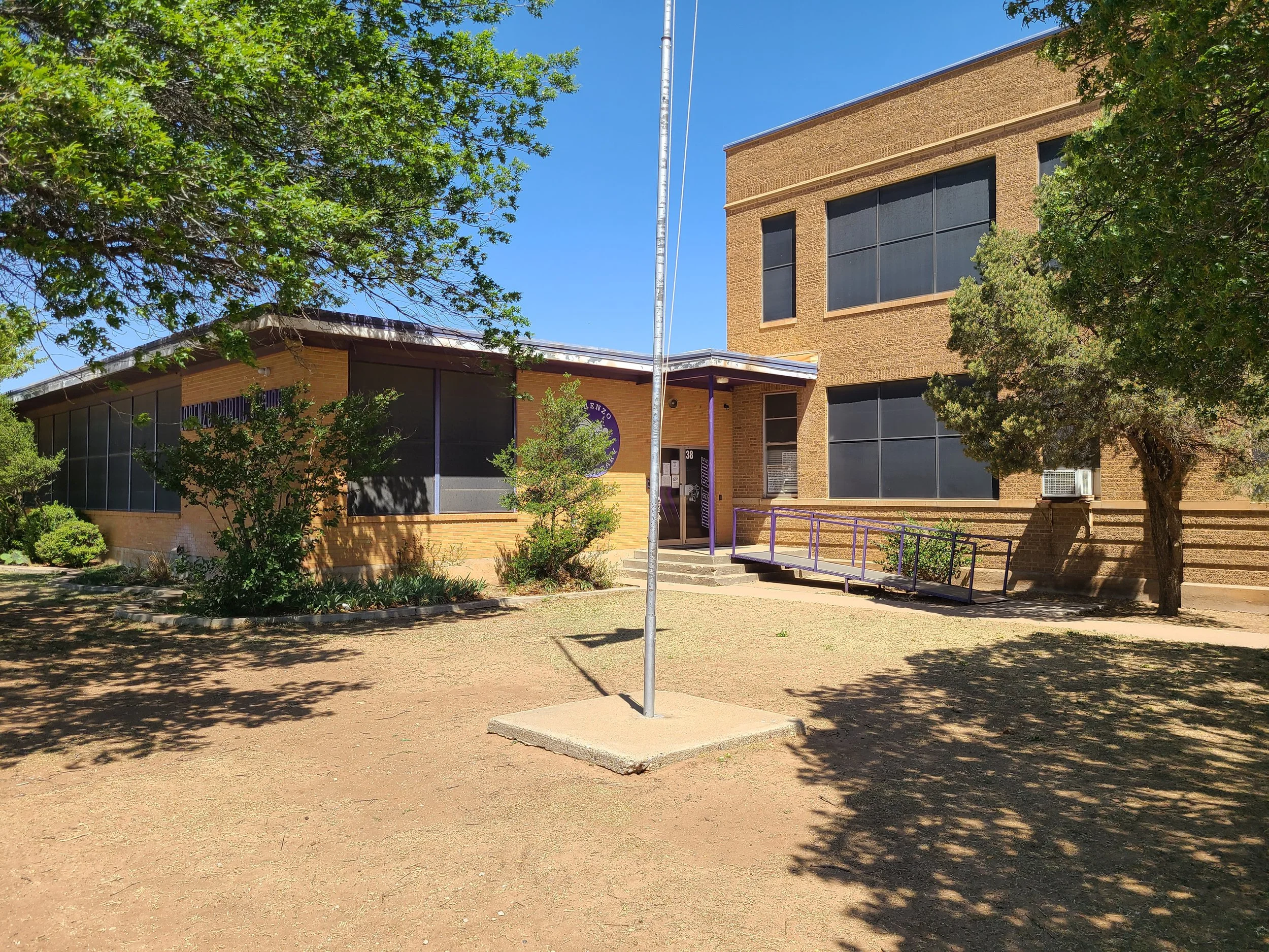 A brick school building with large windows, surrounded by trees and a yard, under a blue sky. A flagpole stands in front of the building.