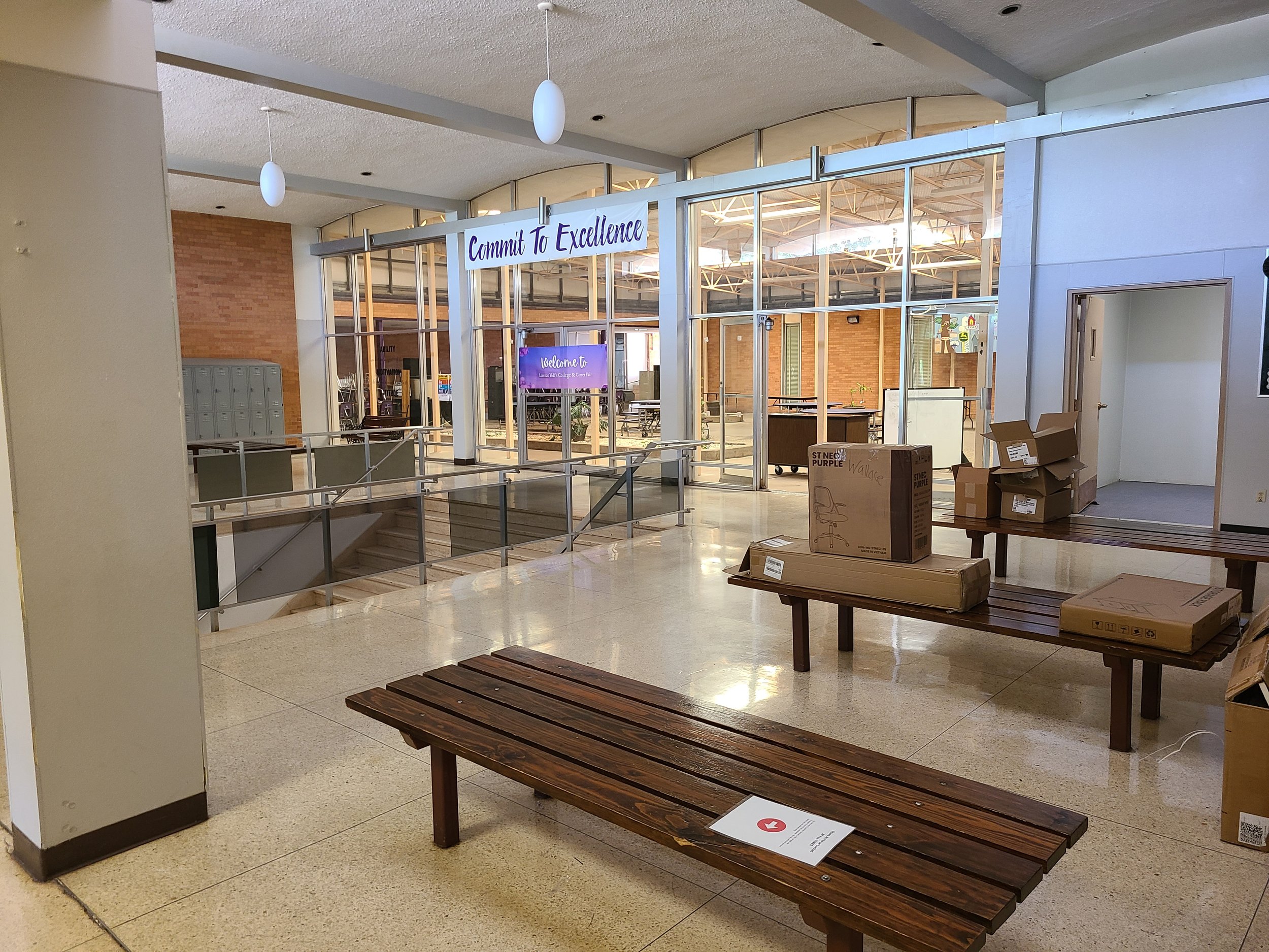 School lobby with lockers, wooden benches, a staircase, and a large entrance with a sign that says "Commit To Excellence." Boxes are stacked on a table near the benches, and a digital display welcomes visitors.