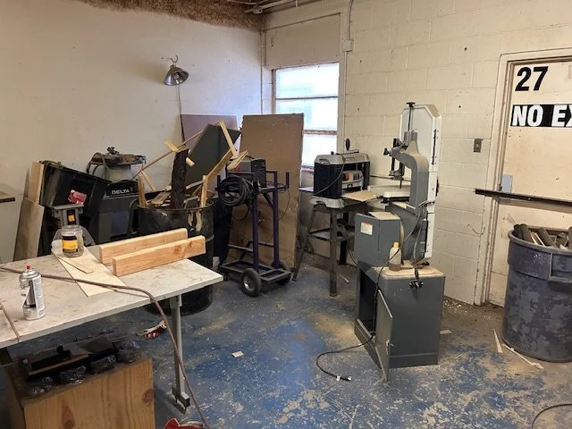 Workshop with various tools, a workbench, wood pieces, and a bandsaw in a cluttered space with a window and a door.