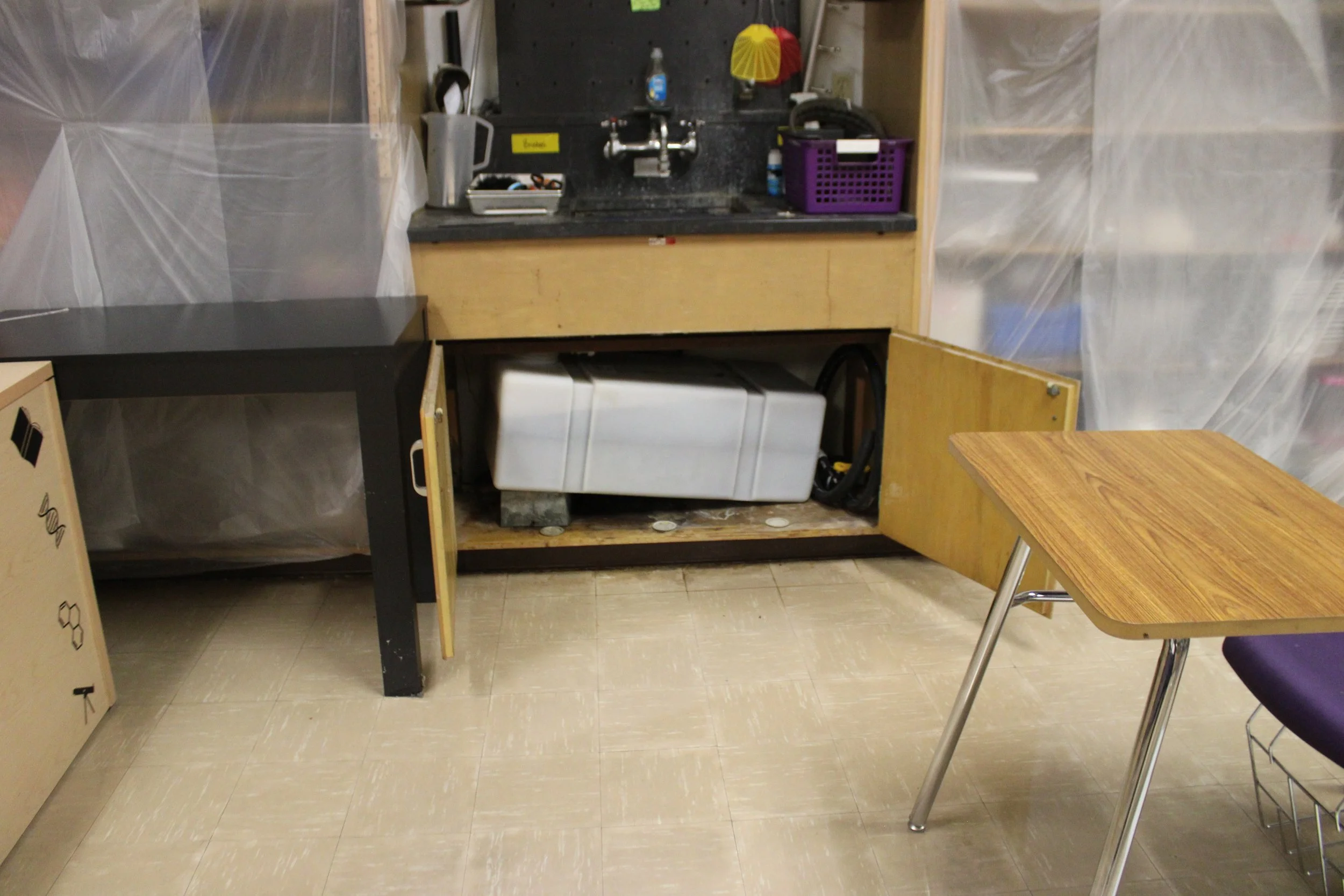 Interior of a small room with unfinished wooden cabinetry and countertops, a plastic storage container underneath, and a small purple basket on the countertop. A folding or portable table is partially visible on the right, and the walls are covered w