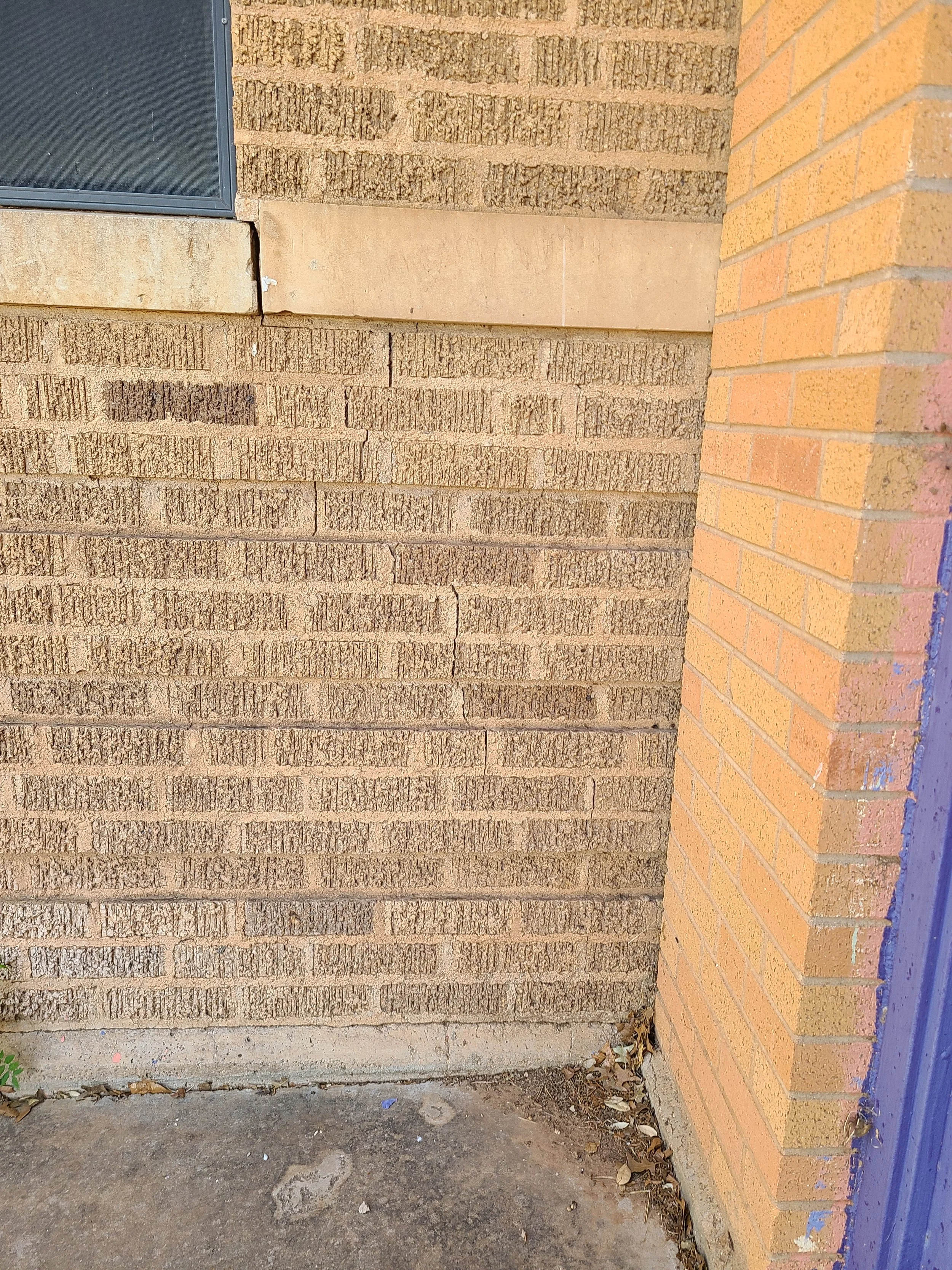 Close-up of a brick wall with a window and a concrete sidewalk at the bottom, with some dirt and leaves on the ground.