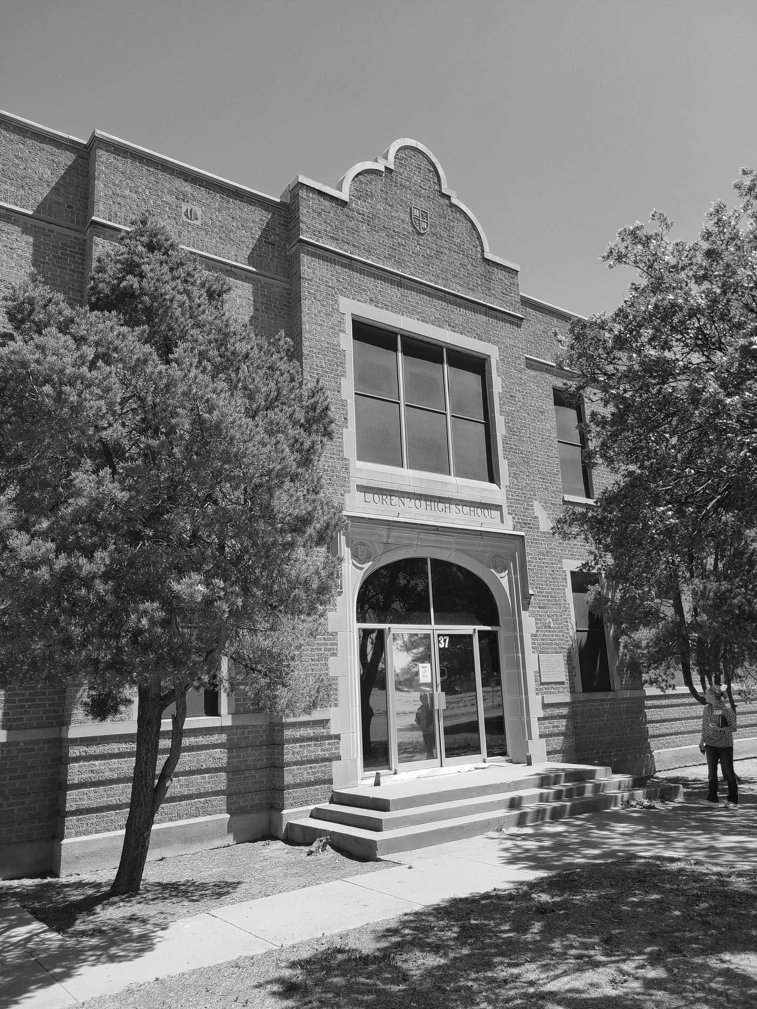 Black and white photo of the front entrance of Lorenzo High School, with steps leading up to a large glass door, trees on either side, and a sign above the door. The school building has brick walls and a shield crest near the top.