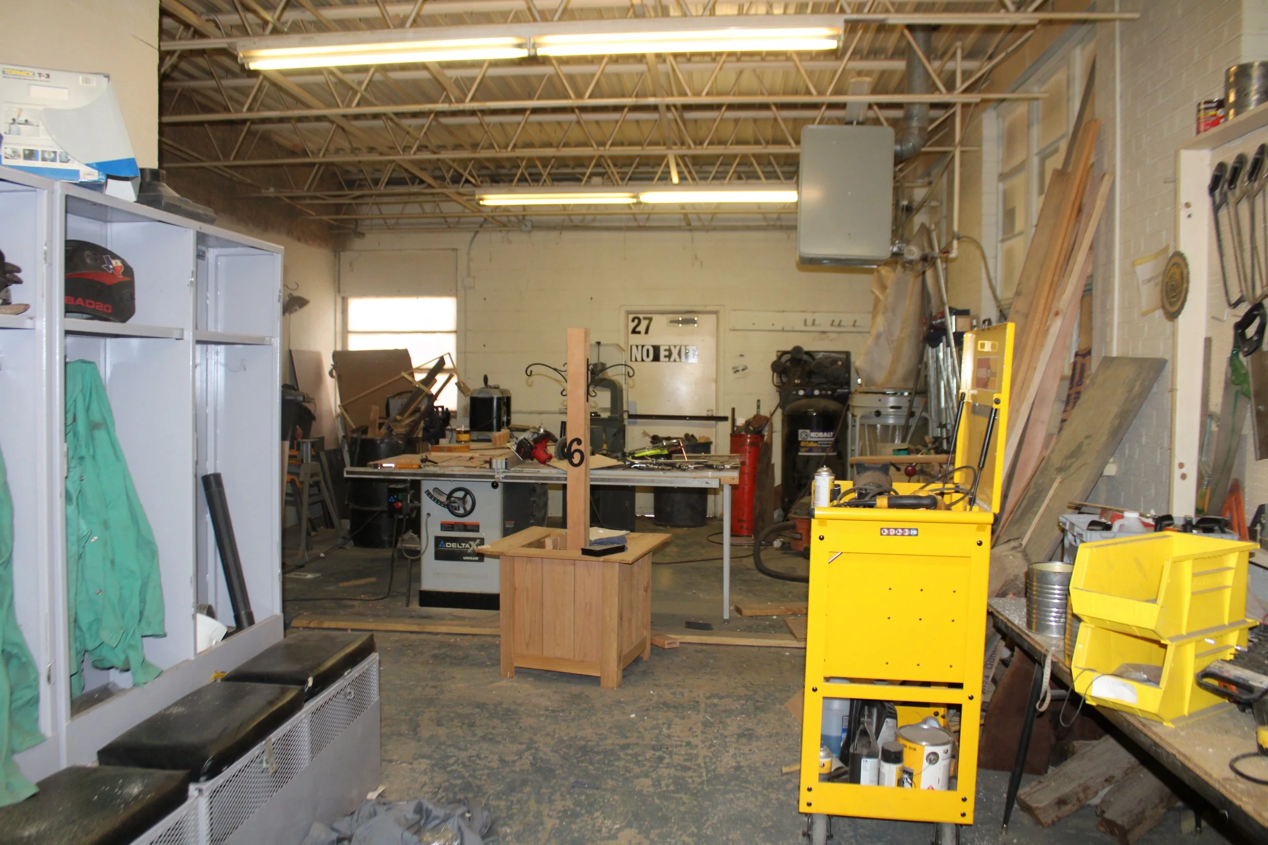 A cluttered workshop with workbenches, tools, and equipment, including a yellow tool chest, lockers, and assorted woodworking supplies, under fluorescent lighting.