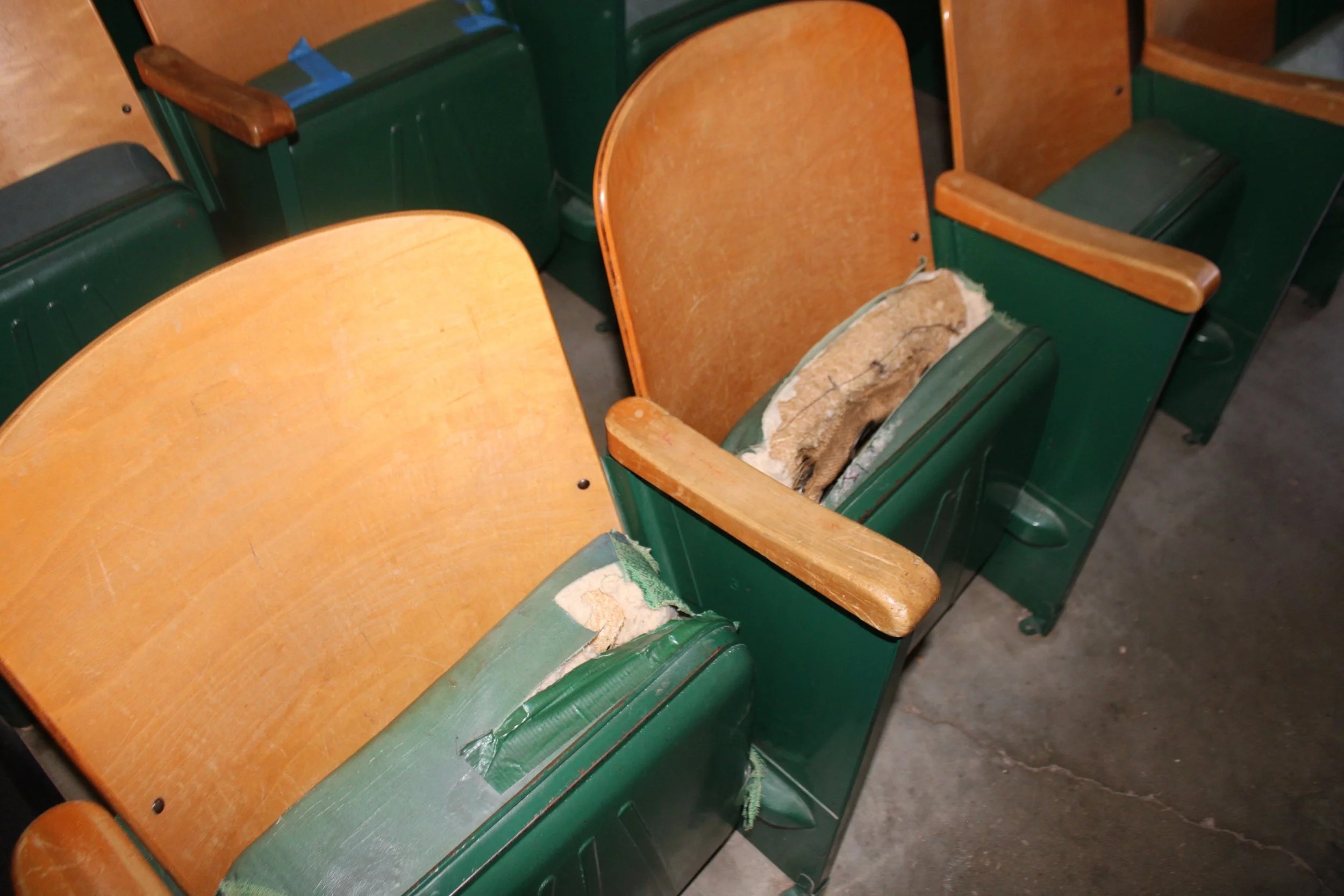 Row of old wooden and green stadium or theater seats, some with torn or worn upholstery.
