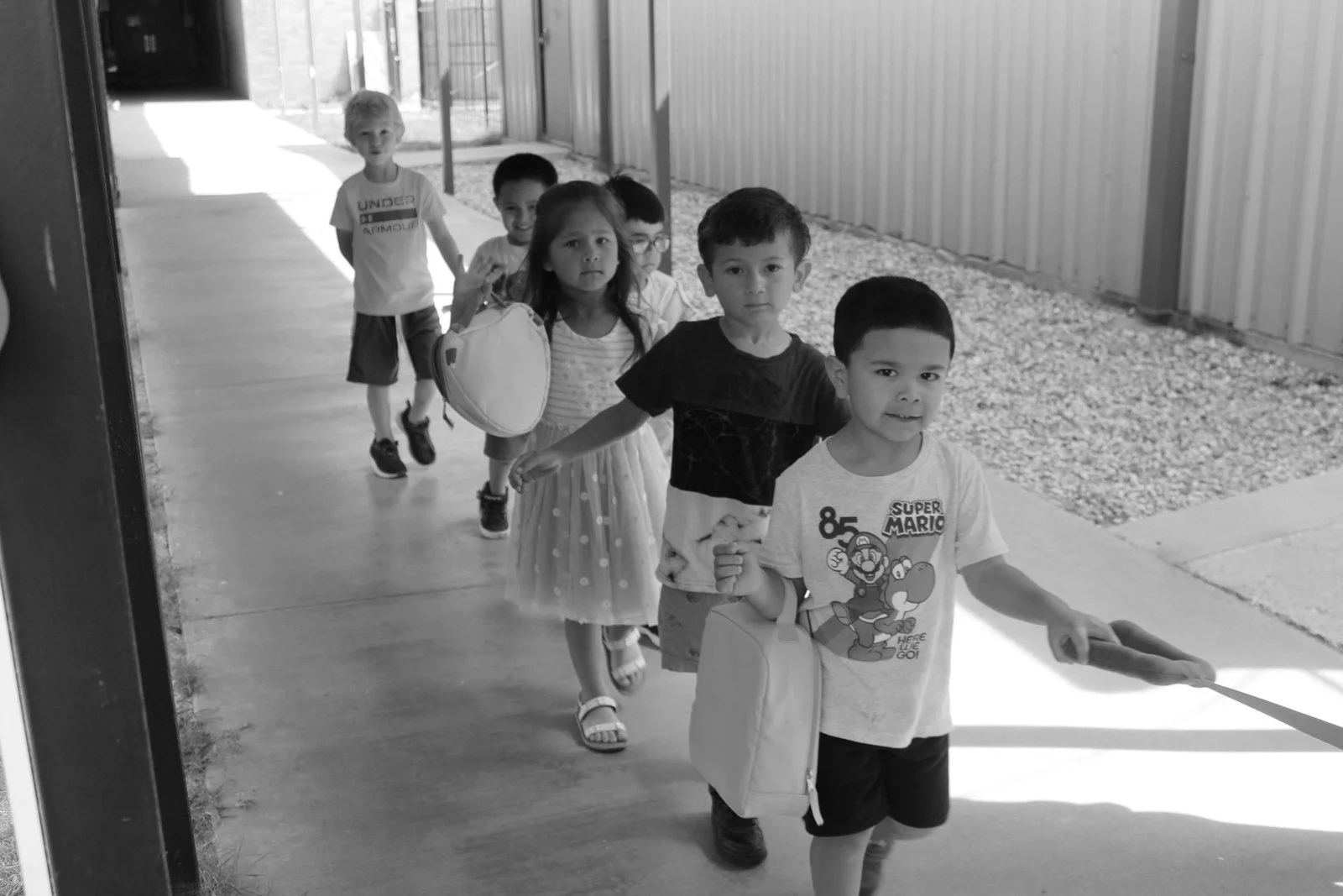 A line of children walking outdoors on a sidewalk, with a boy in a Super Mario T-shirt at the front holding a bag and a stick, followed by other kids, some carrying backpacks and holding hands.