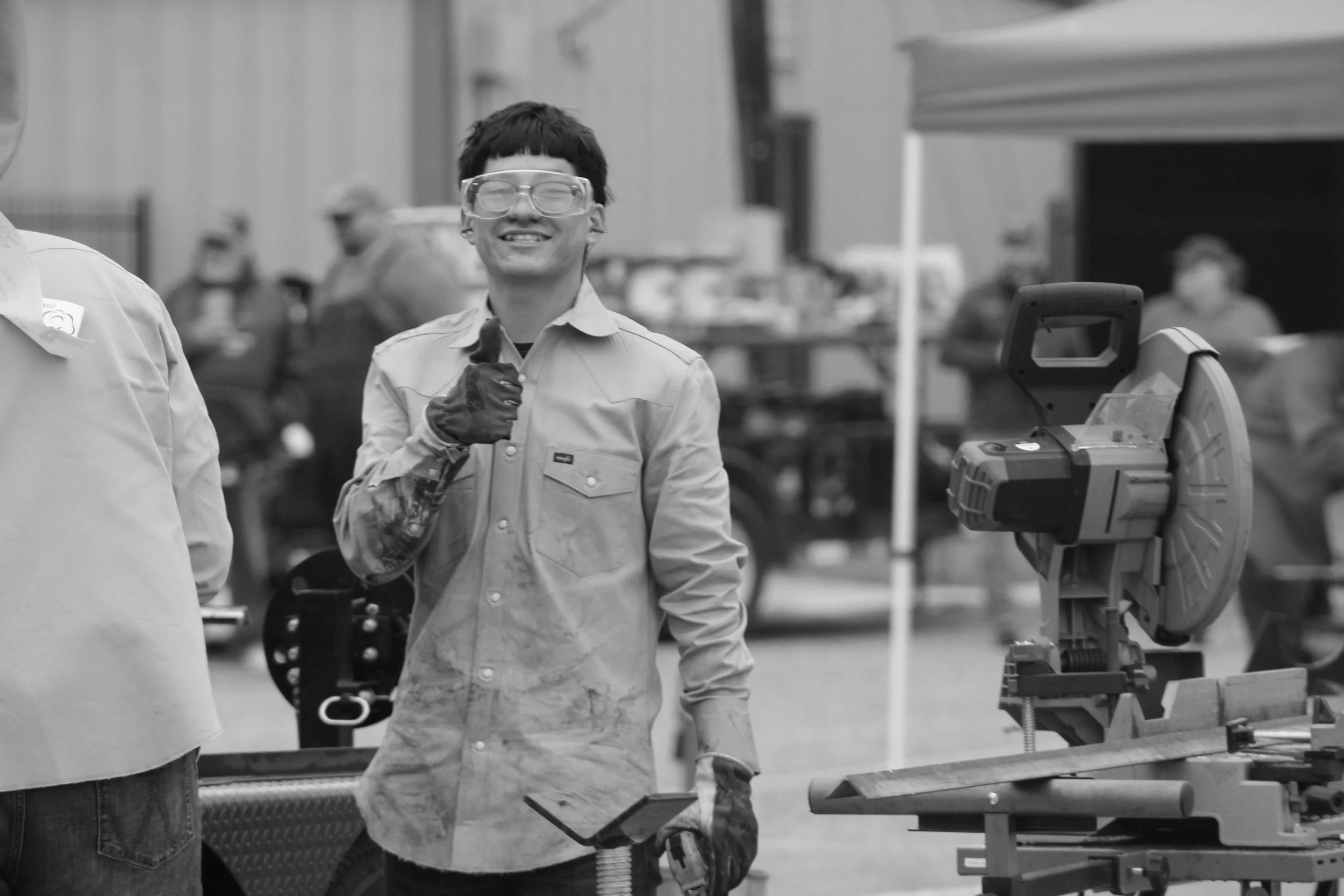 A smiling young man wearing safety glasses and work gloves, standing near a workbench and power tools in an industrial or workshop setting.